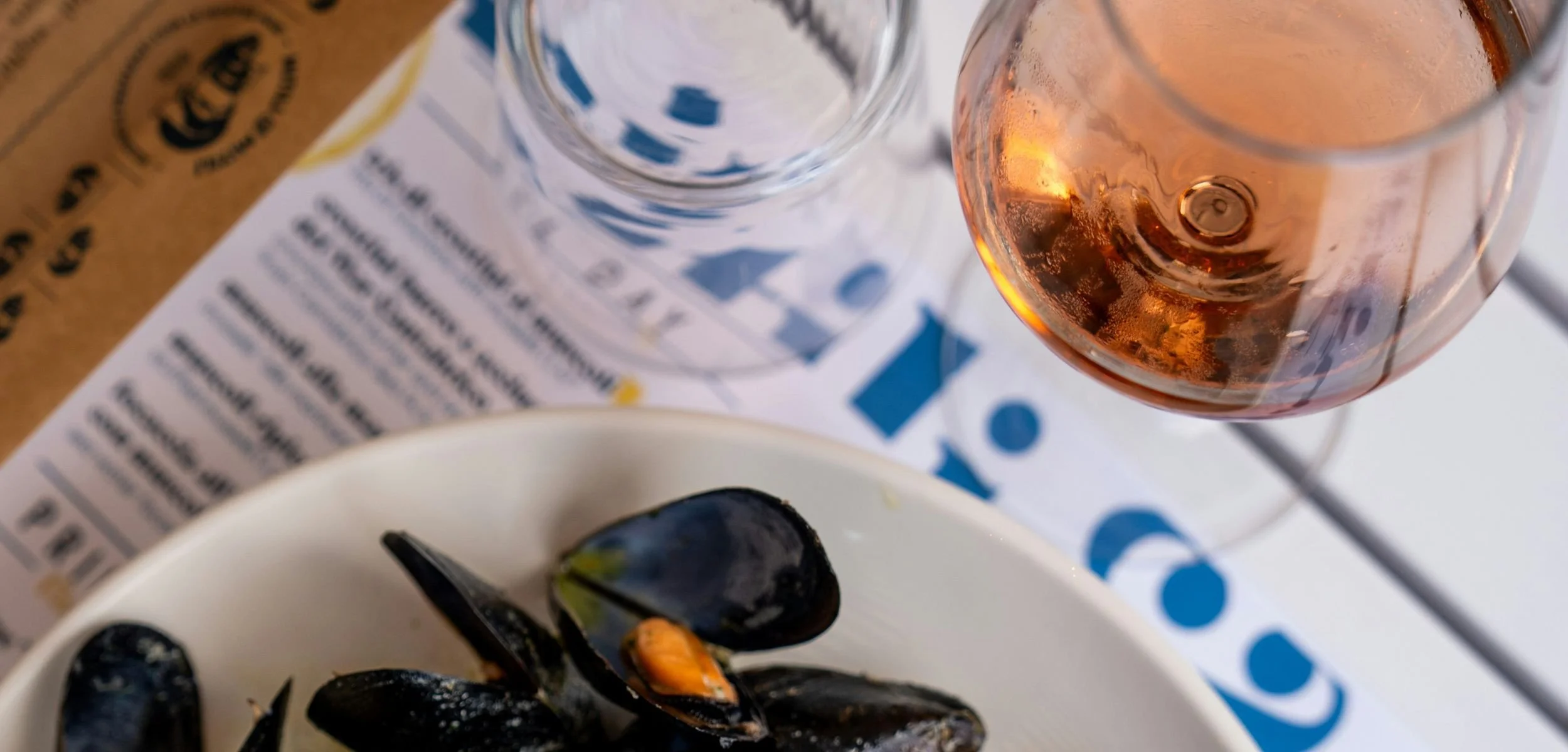 A bowl of fresh black mussels served next to a glass of chilled non-alcoholic rose wine on a white table with a printed menu and a clear drinking glass nearby.