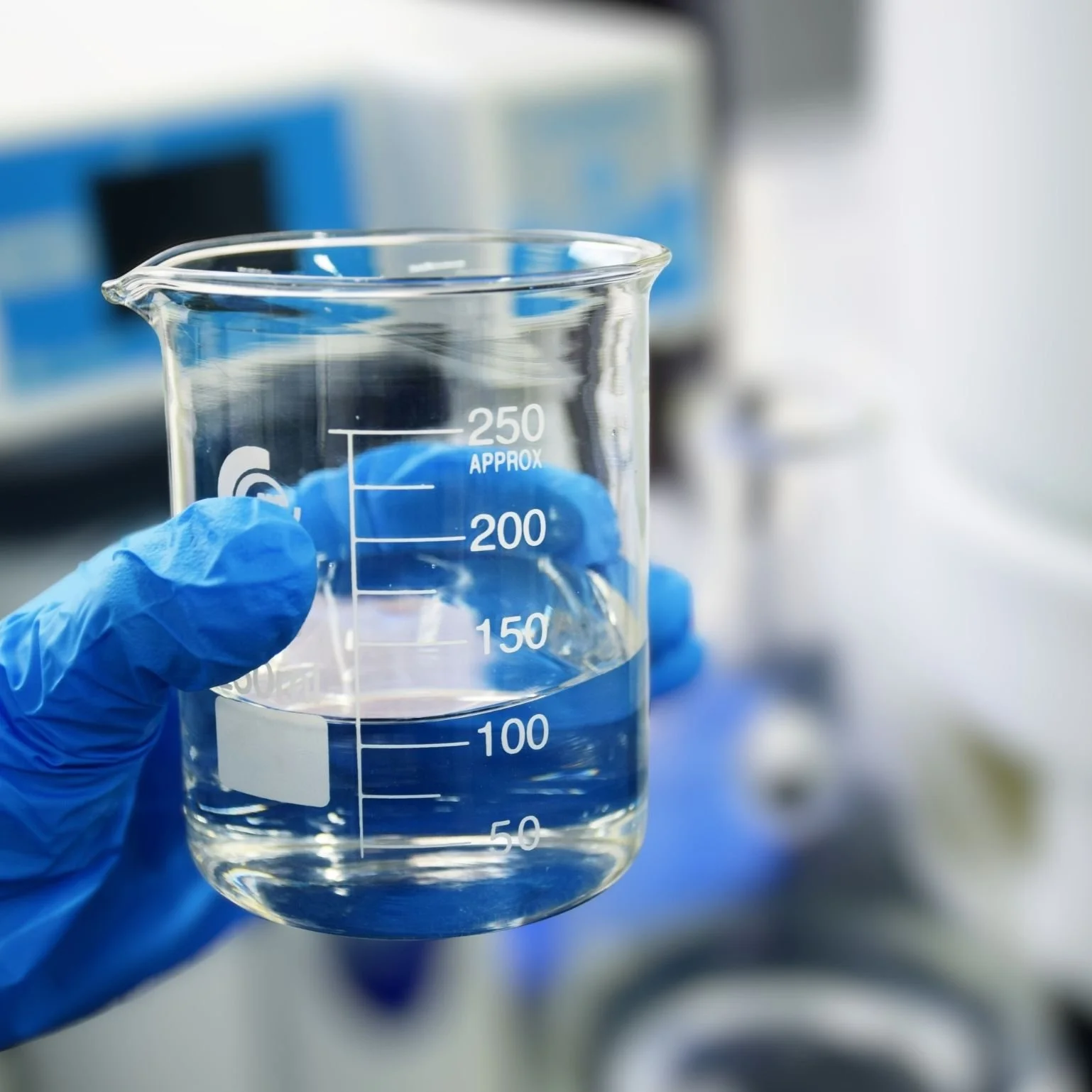 A hand wearing a blue glove holds a glass beaker filled with liquid, marked with measurement lines.