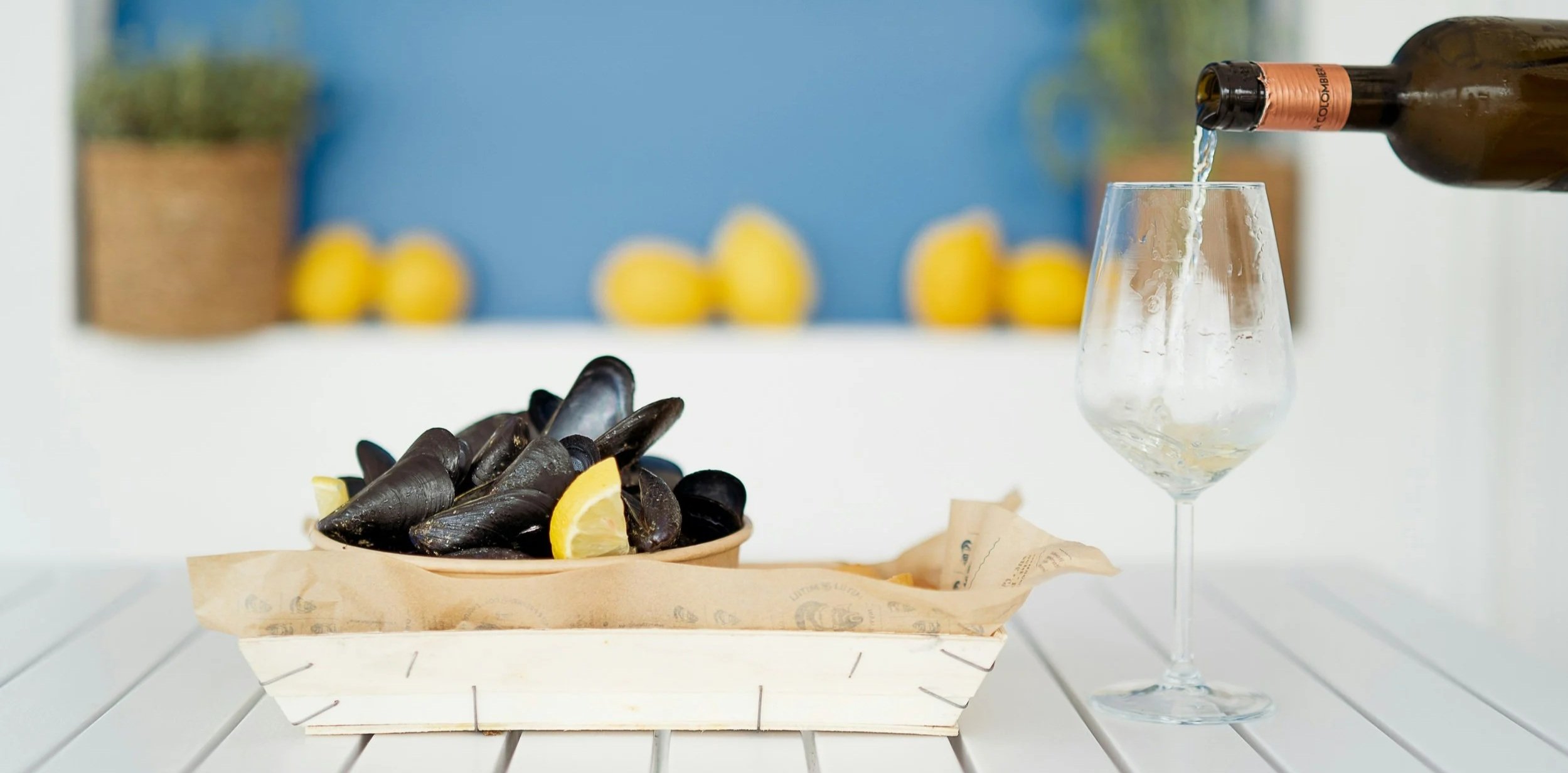 A bowl of fresh black mussels with lemon wedges in a wooden crate next to a glass being filled with non-alcoholic white wine against a blue and white background.