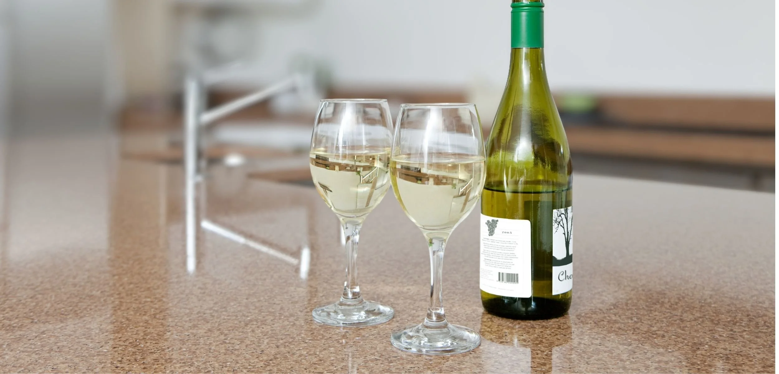 A bottle of de-alcoholized white wine and two filled wine glasses arranged on a stone countertop in a bright kitchen setting.