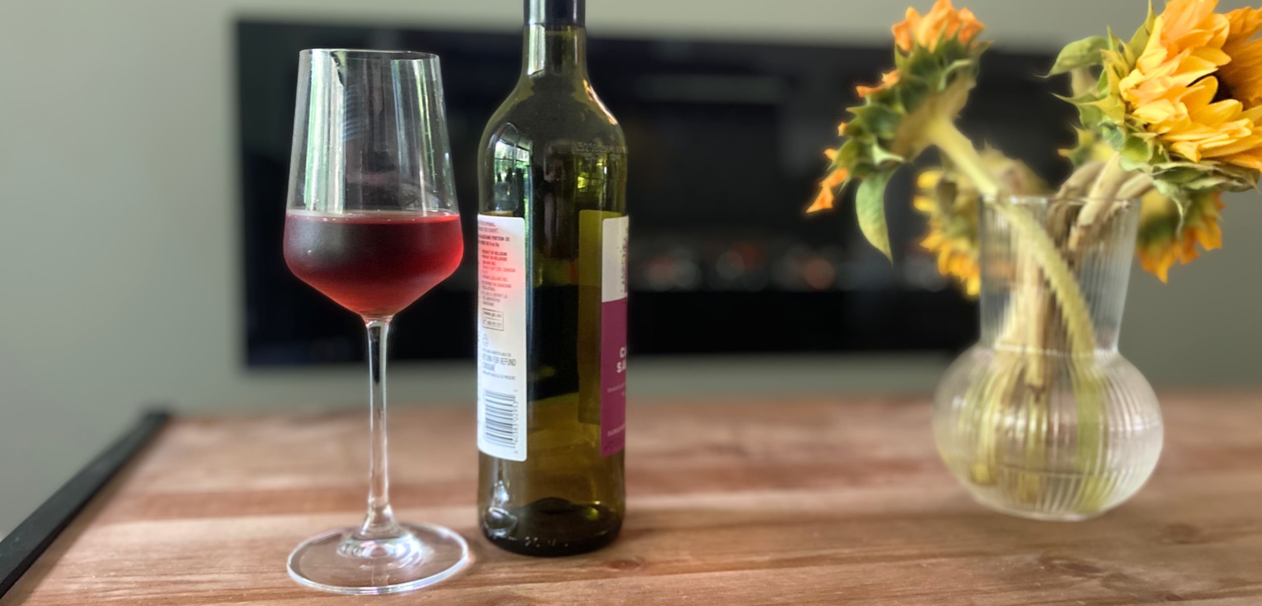 Non-alcoholic red wine in a stemmed glass and a bottle arranged on a wooden table, with yellow sunflowers in a vase and a blurred indoor background.