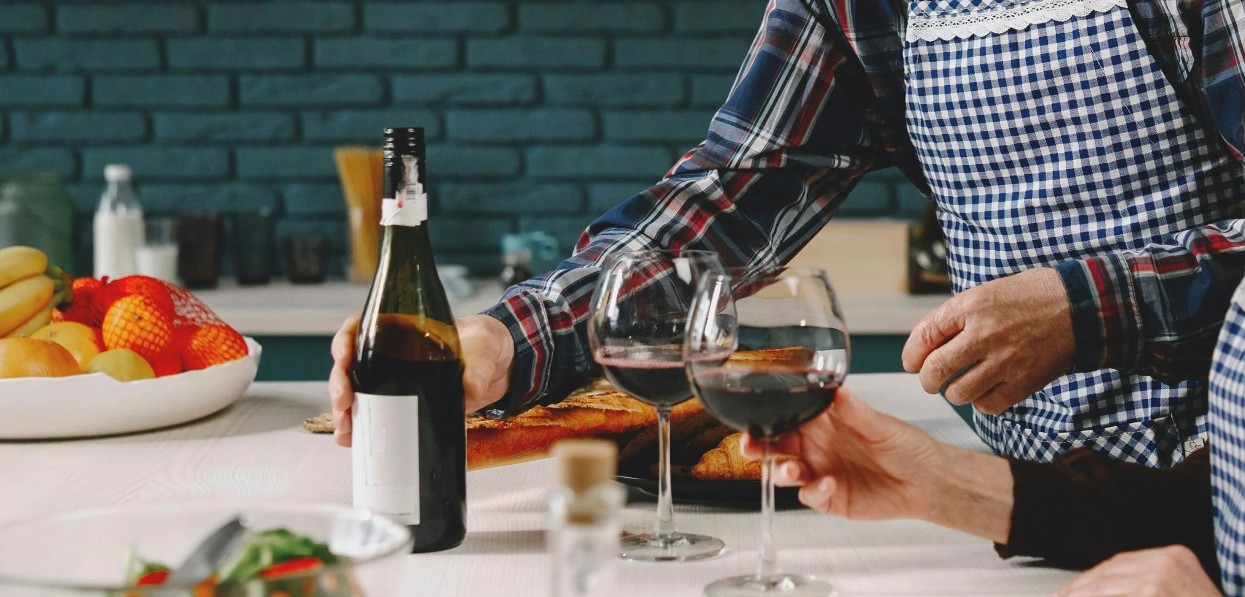 Person in a plaid shirt and apron pours non-alcoholic red wine into a glass held by another person at a table with fruit, bread, and a salad in a home kitchen.