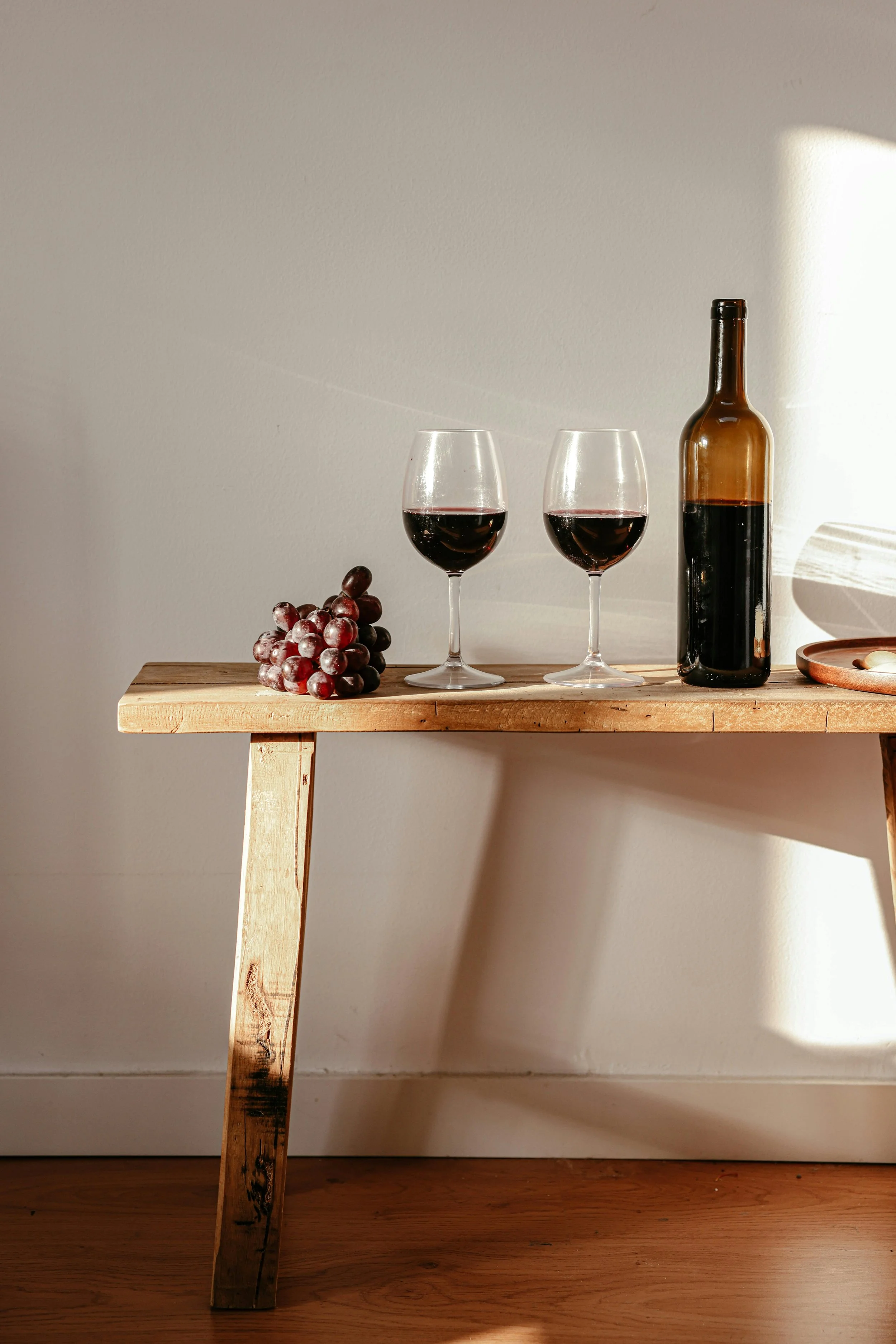 A wooden table with two glasses of non-alcoholic red wine, a bottle, and a bunch of grapes.
