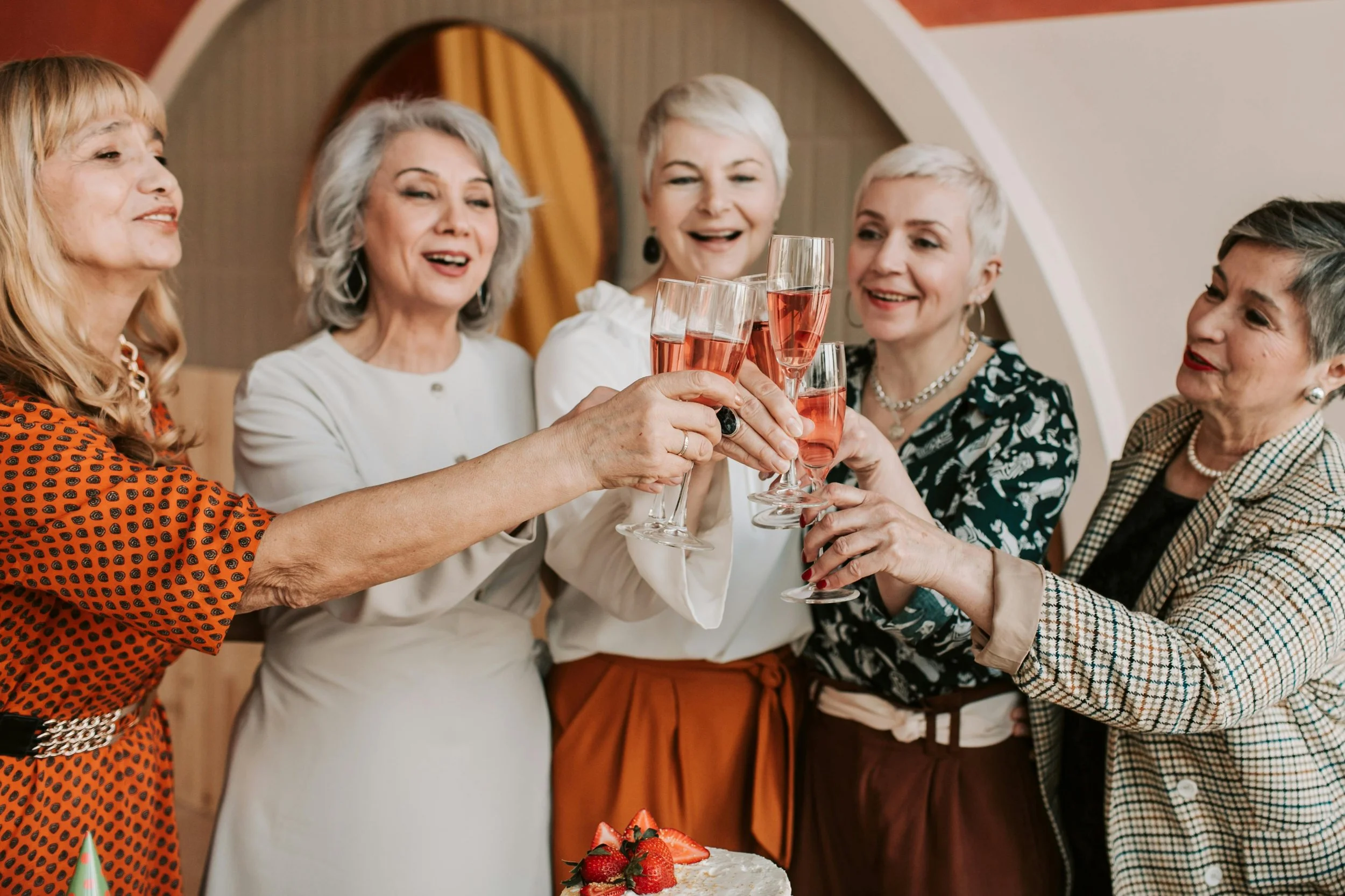 Five stylish women with varying hairstyles and outfits are gathered around a cake, joyfully toasting with champagne flutes.