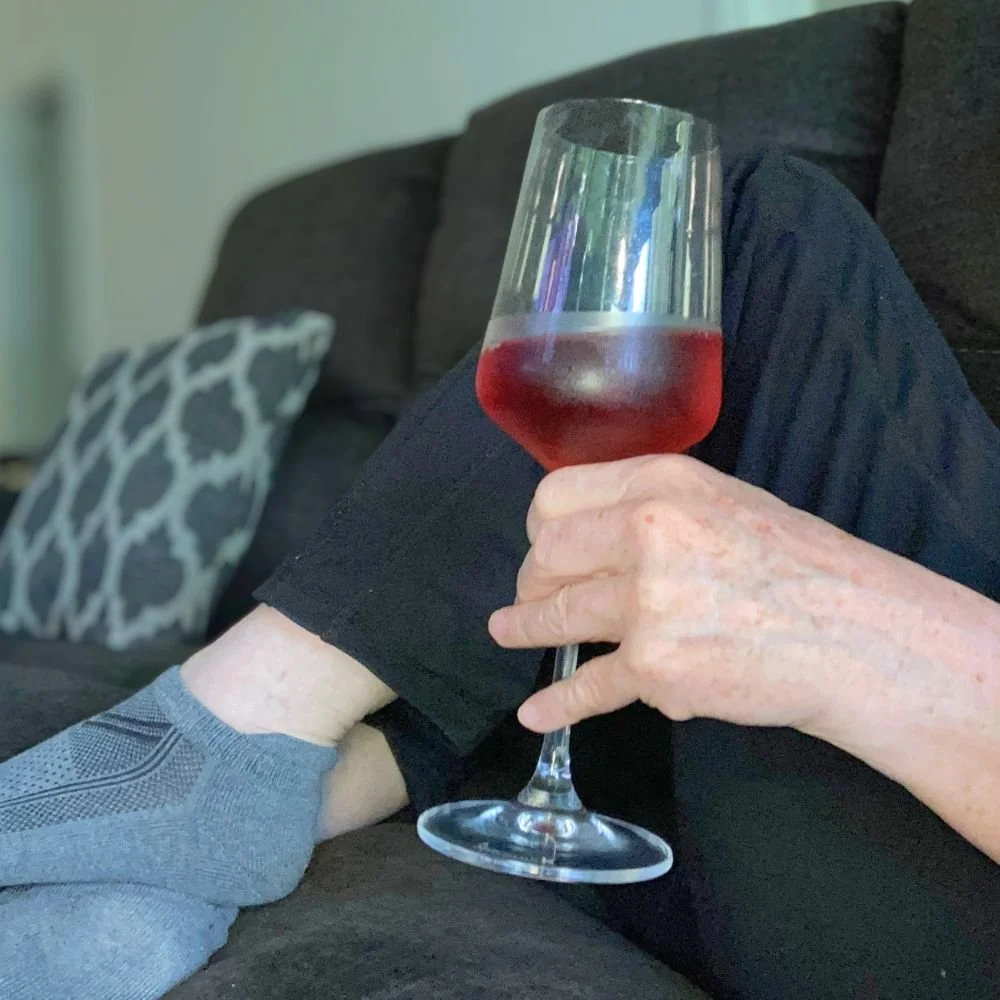 A woman lounging on a couch with a glass of non-alcoholic red wine in hand, creating a cozy indoor scene with soft furnishings in the background.