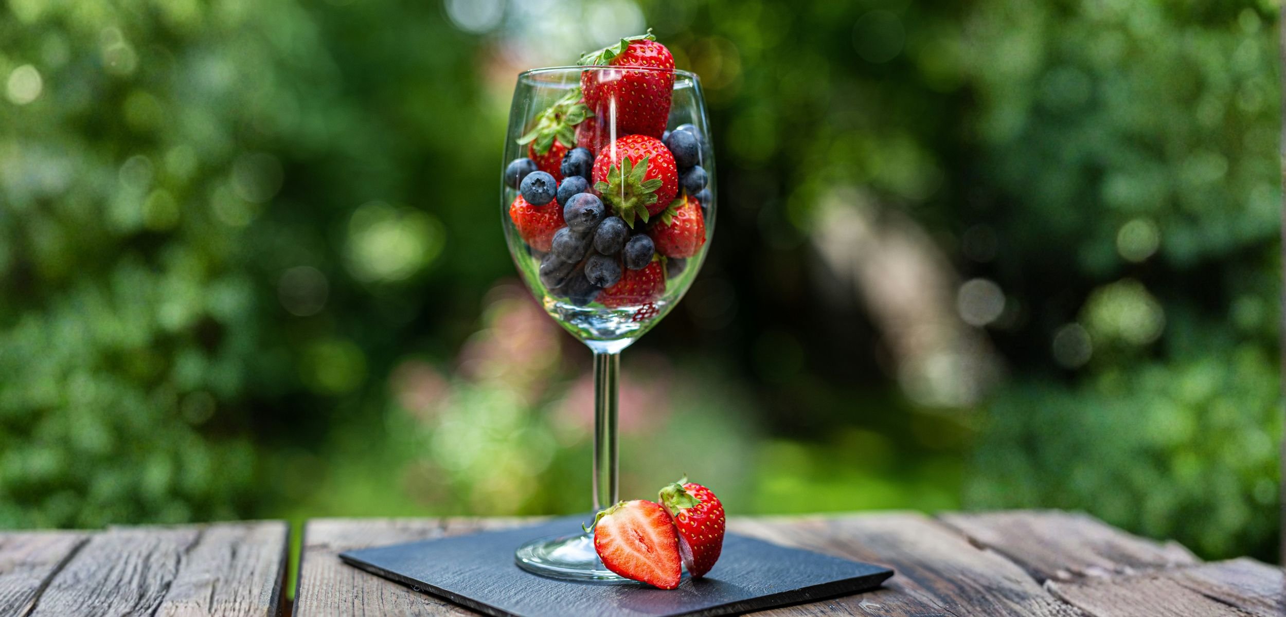 A glass filled with fresh strawberries and blueberries, set on a wooden table in a lush green garden background.