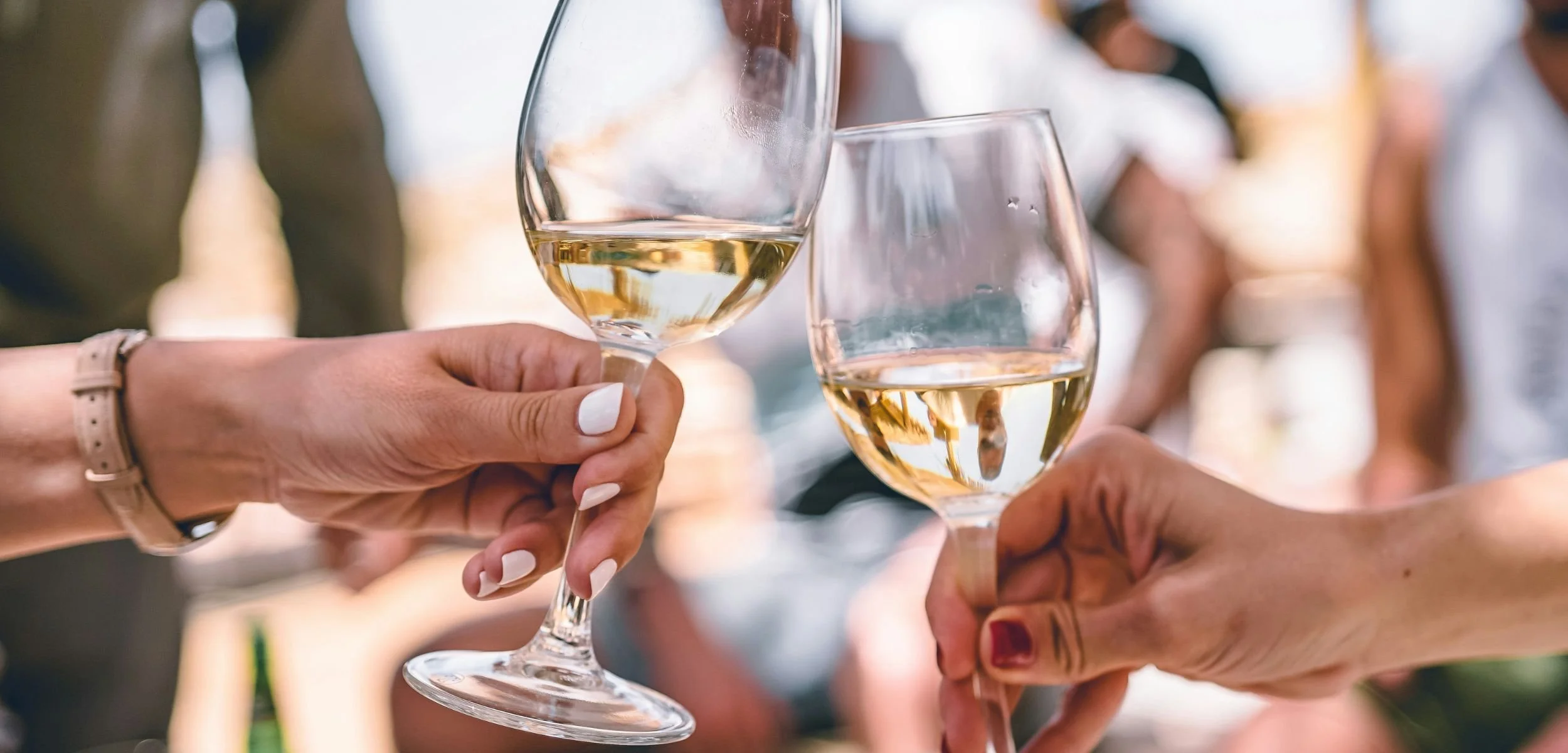Two hands clinking glasses of de-alcoholized white wine in a celebratory toast at a social gathering outdoors.