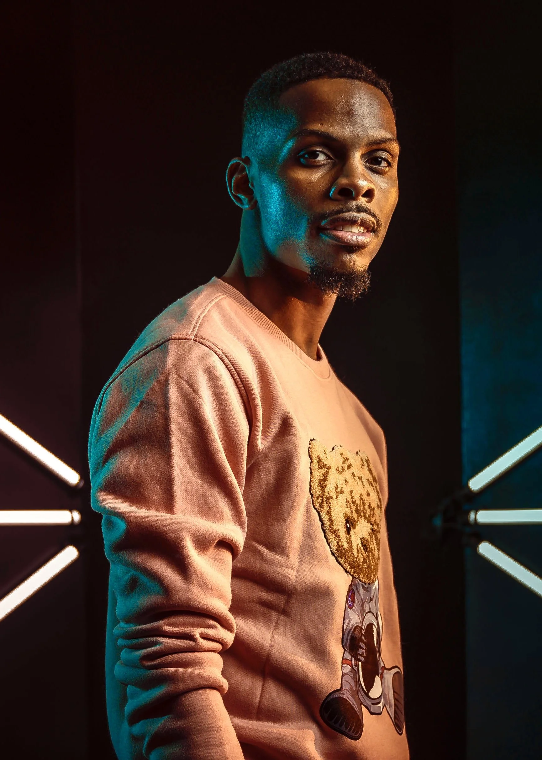 A DJ with short black hair and a goatee, wearing a pink sweatshirt featuring a cartoon character, poses against a dark background with neon lights.