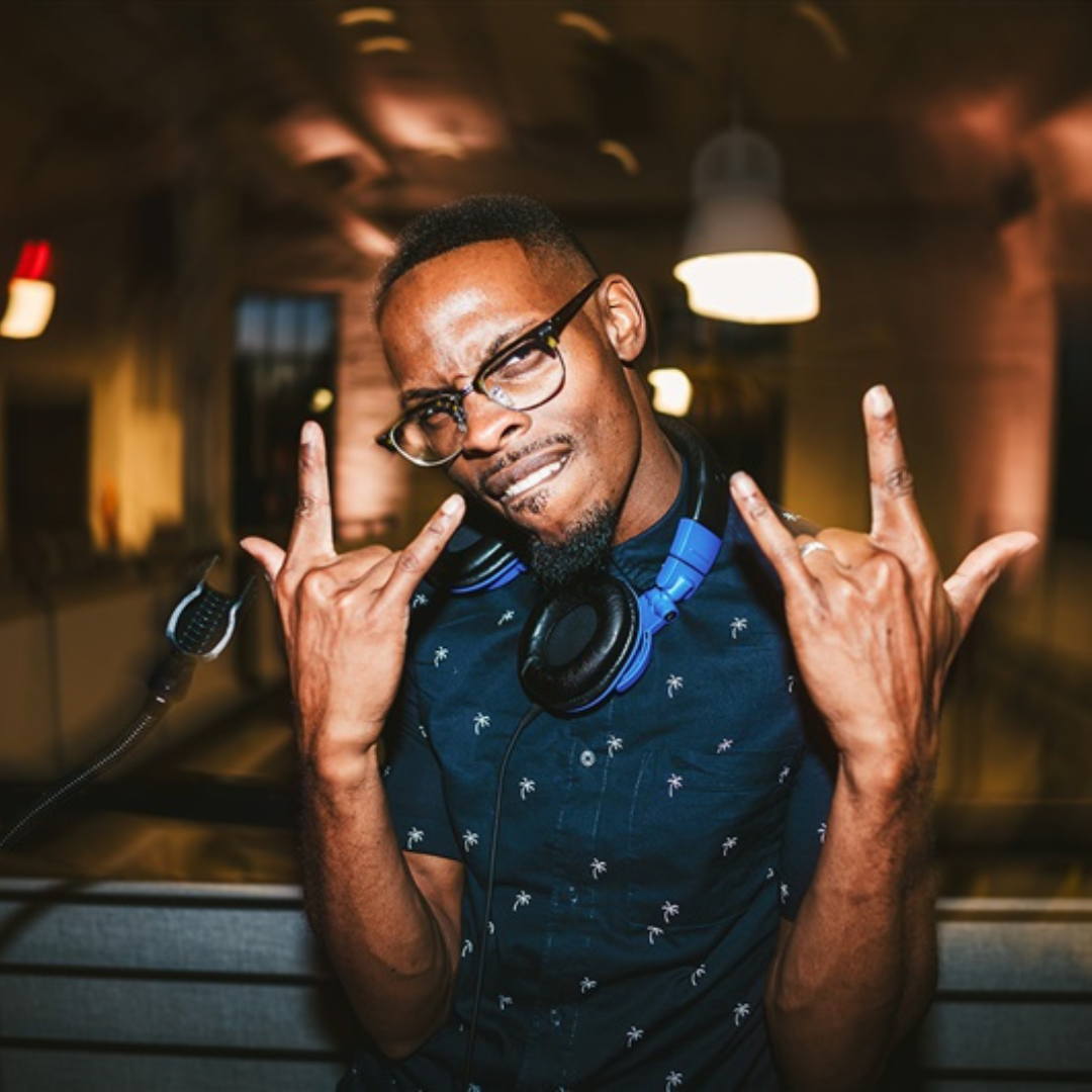 A DJ with glasses and headphones, making rock hand gestures, posing in a dimly lit indoor setting, likely a nightclub or music event.