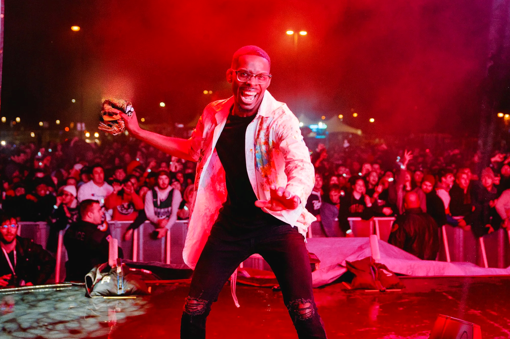 A DJ dancing confidently on stage at a crowded concert, illuminated by red stage lighting, with an audience in the background.