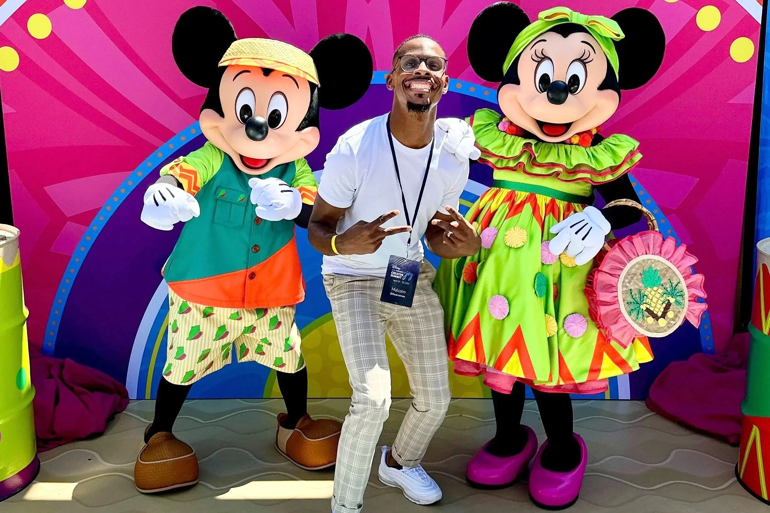 A DJ in white checkered pants and a white t-shirt is posing with two people dressed as Minnie Mouse characters in colorful outfits, against a pink animated backdrop.