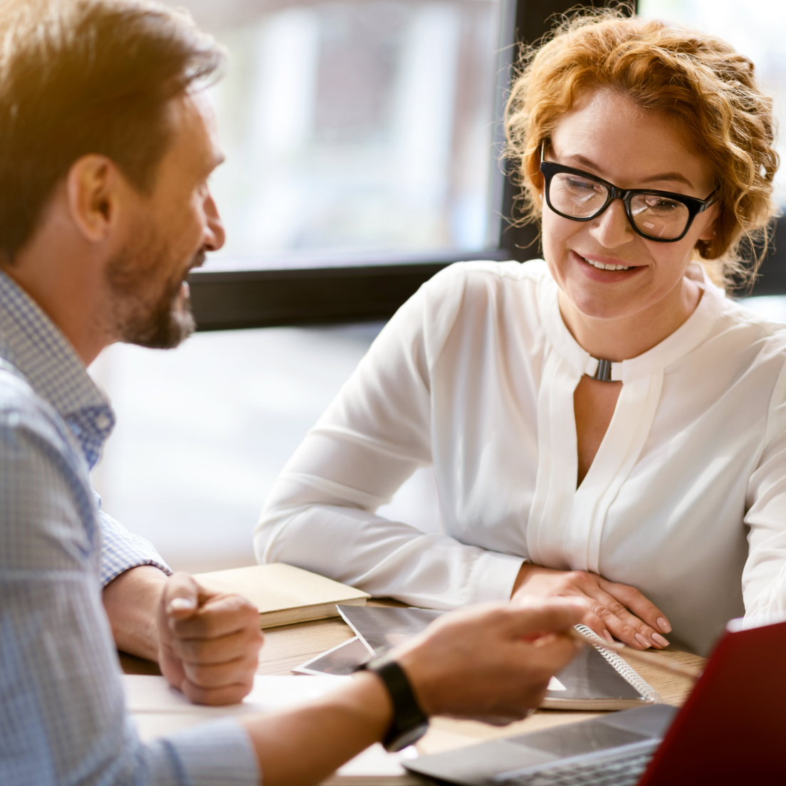 A woman with curly red hair and glasses smiling while talking to a man with brown hair, both sitting at a table with notebooks and a laptop in a bright indoor setting.
