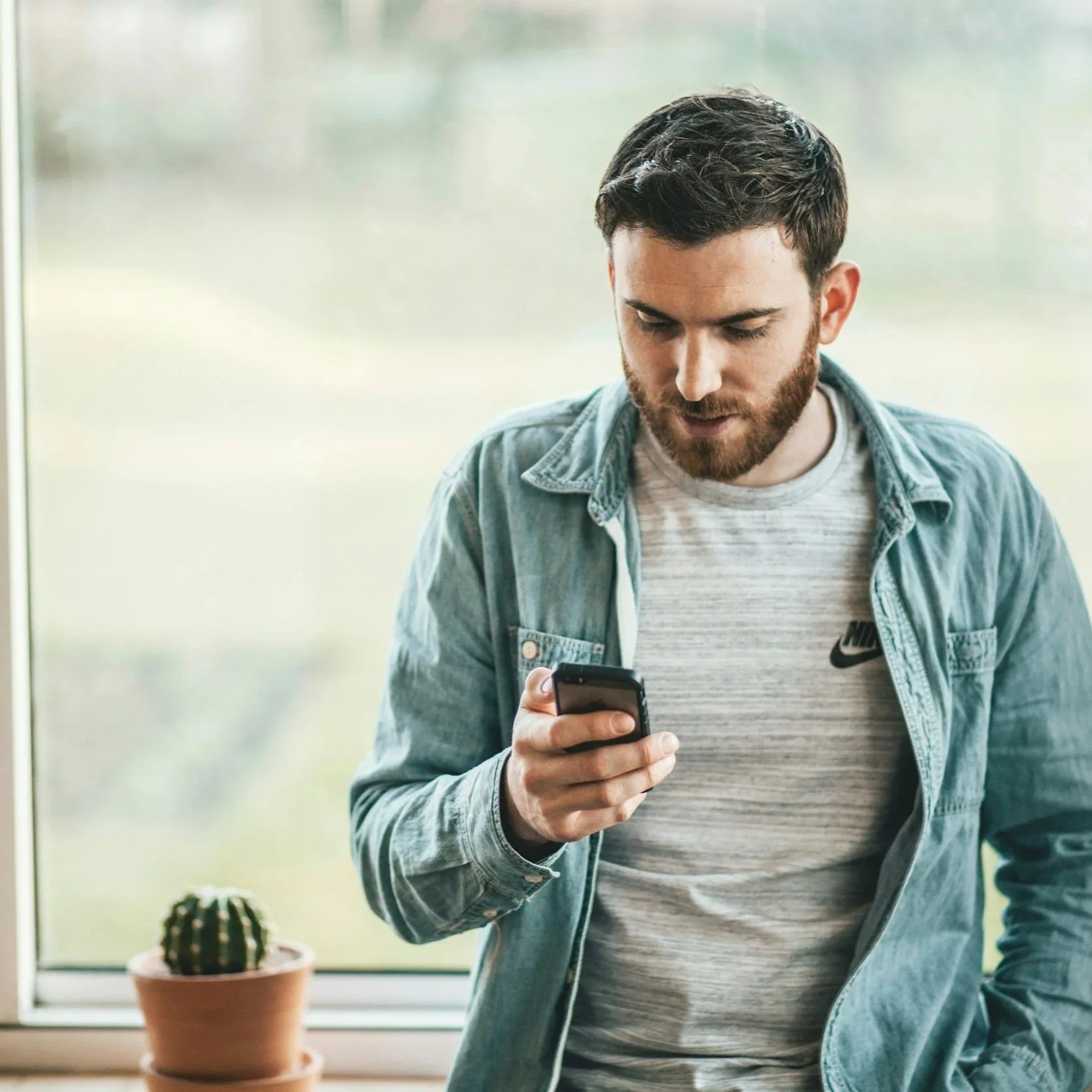 A man with a beard and dark hair wearing a light gray Nike t-shirt and denim jacket looking at his smartphone, with a potted cactus plant on the windowsill nearby.