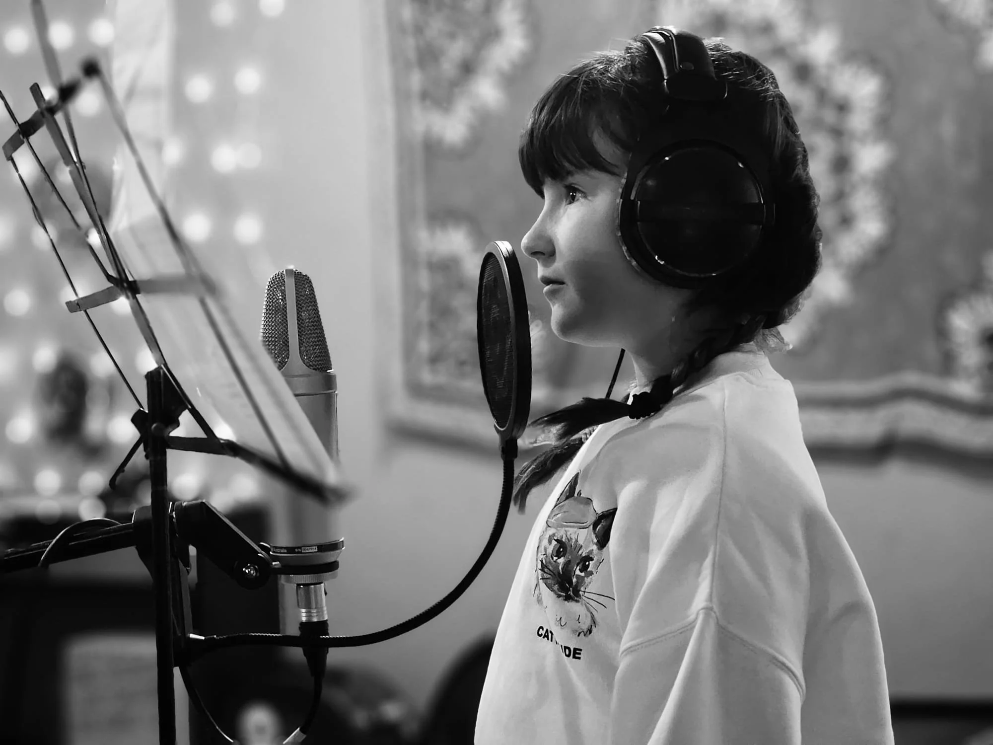 A young girl in a sweatshirt with a cat design, wearing large headphones, standing in front of a microphone in a recording studio.