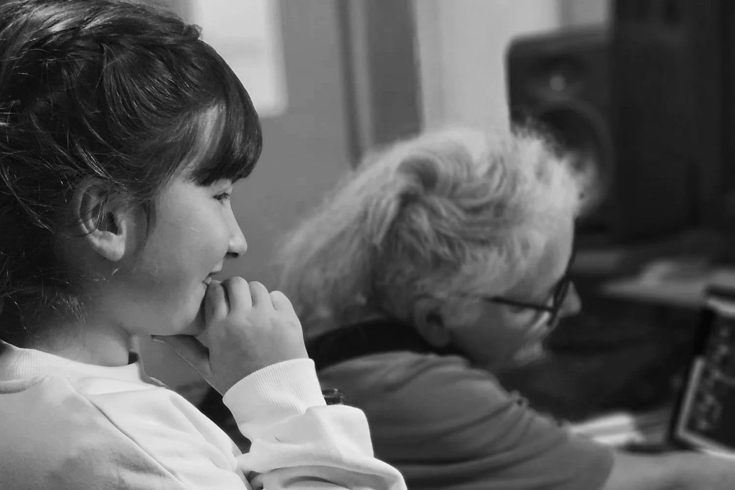A young girl with dark hair and a woman with white hair and glasses sitting in front of a computer or television screen.