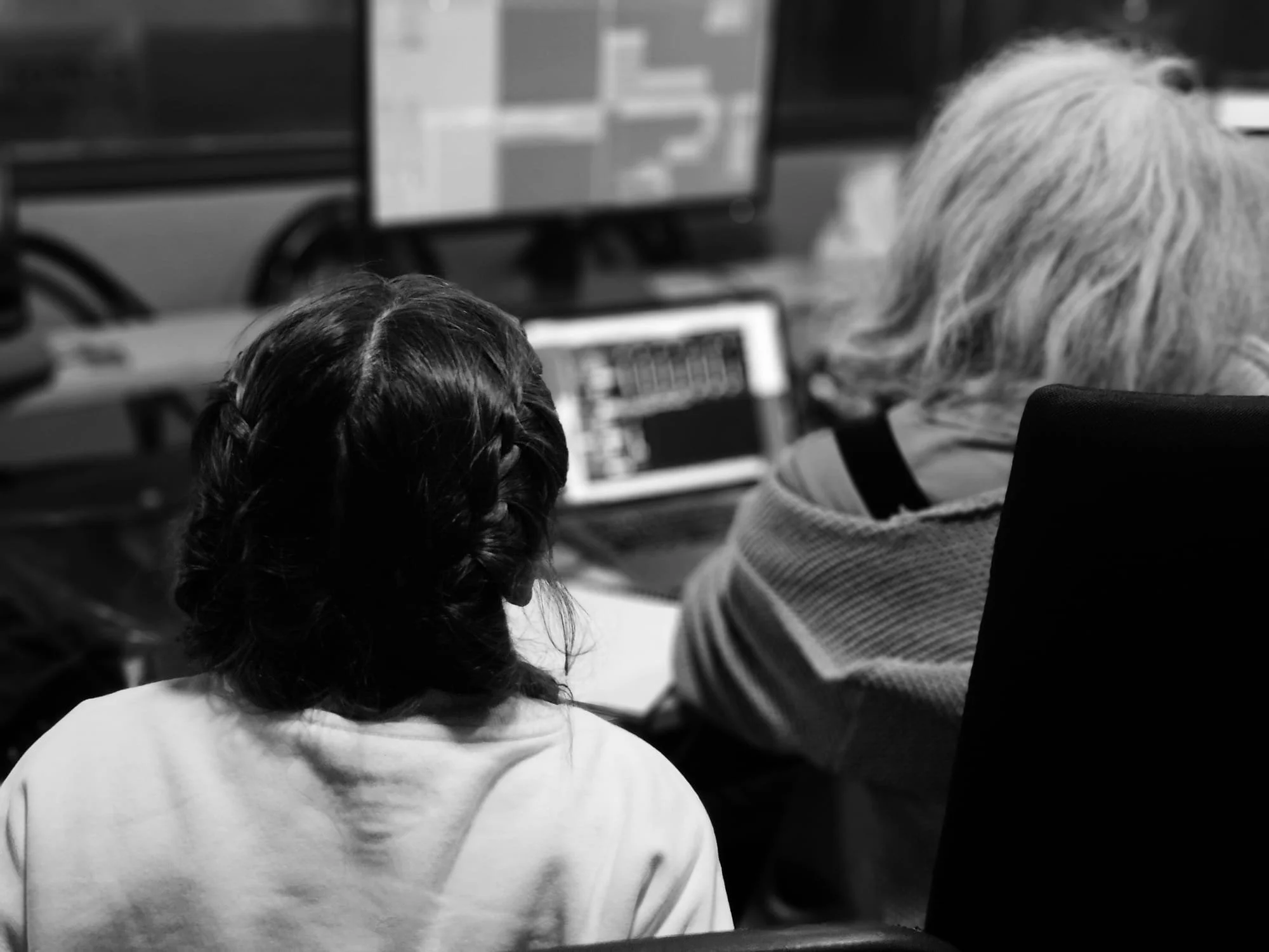 Two women, one with braided dark hair and another with gray hair, sitting at a desk working on computers in an office setting.