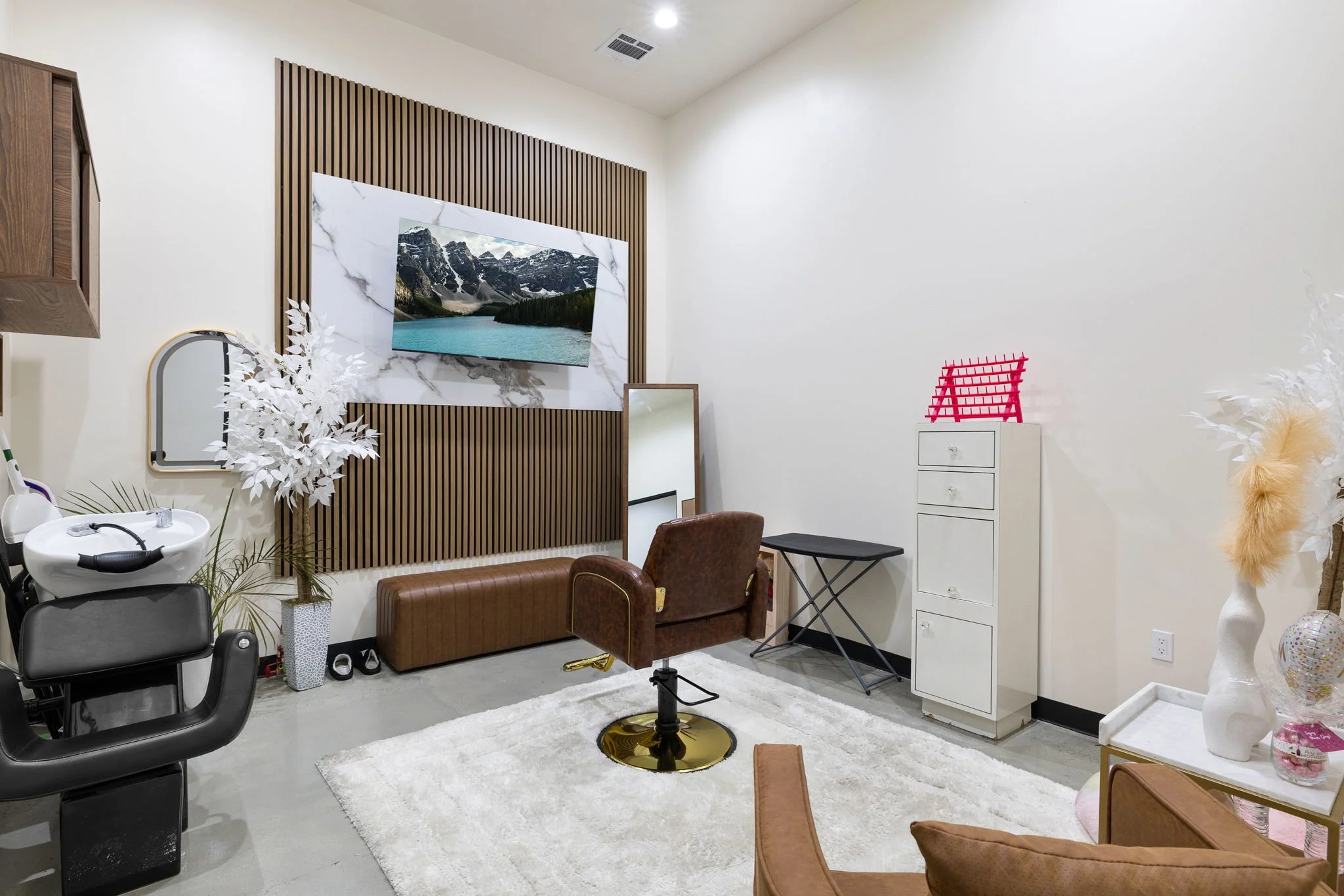 Interior of a room with a brown salon chair, a mirror, a white cabinet with drawers, a white blanket, and decorative vases and plants. A mountain landscape TV is mounted on the wall with a wooden slat wall behind it.