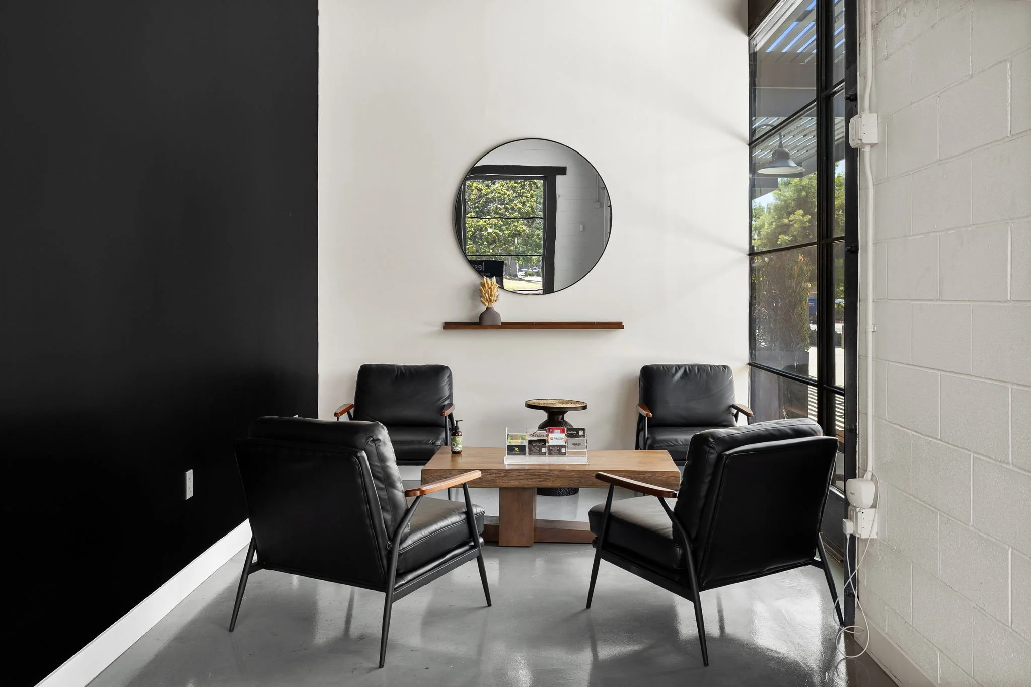 A modern waiting or meeting area with four black leather armchairs arranged around a wooden coffee table, decorative items on the table, a round mirror on a white wall, a wooden shelf with a vase, and large glass windows letting in natural light.