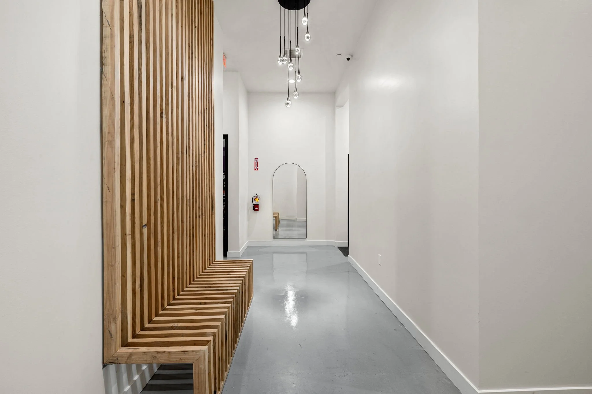 Empty hallway with wooden slat seating on the left, white walls, gray flooring, mirror, fire extinguisher, and modern pendant light fixtures