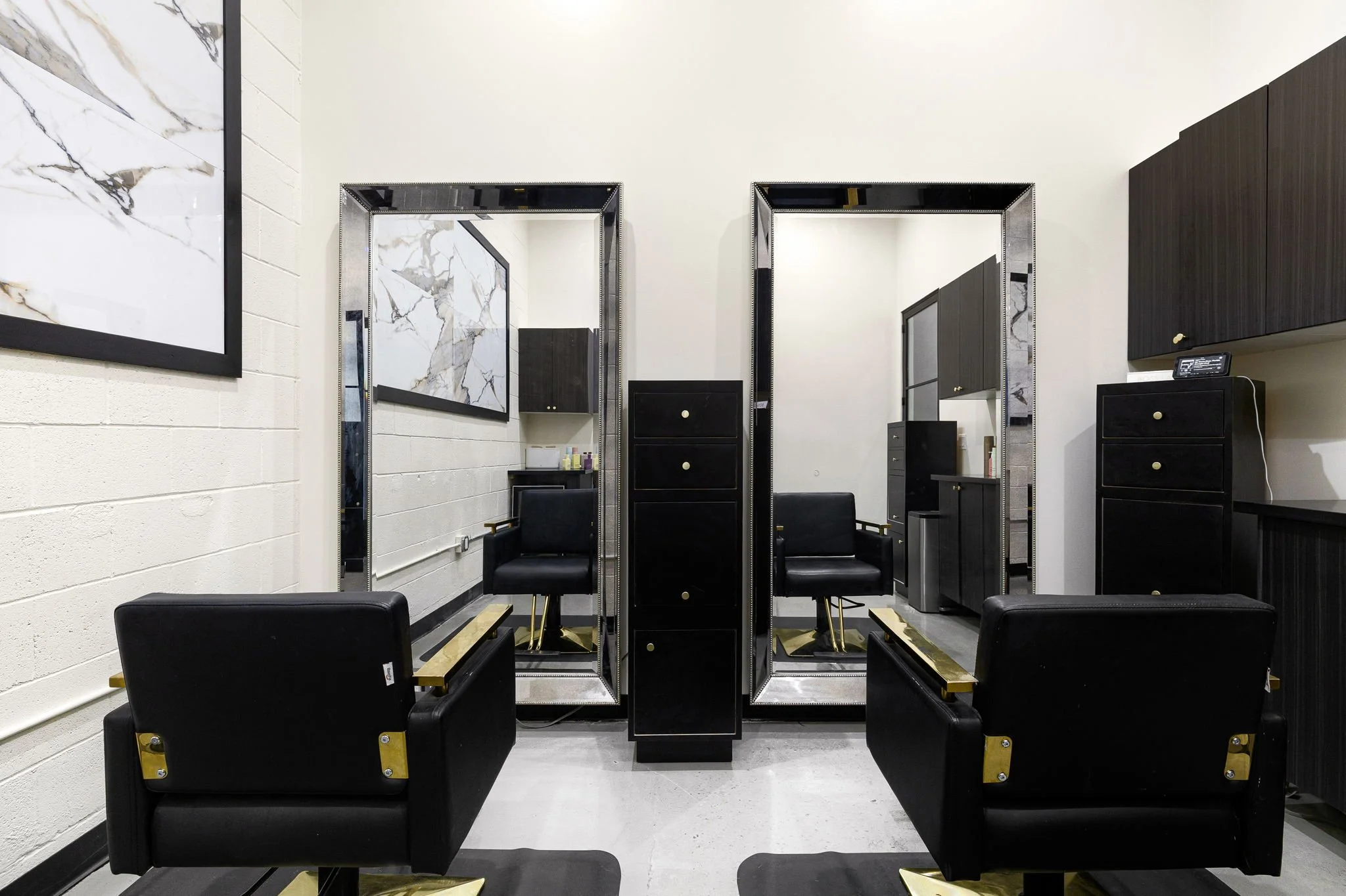 Two salon chairs facing large mirrors in a modern hair salon with black and gold decor and artwork on the wall.