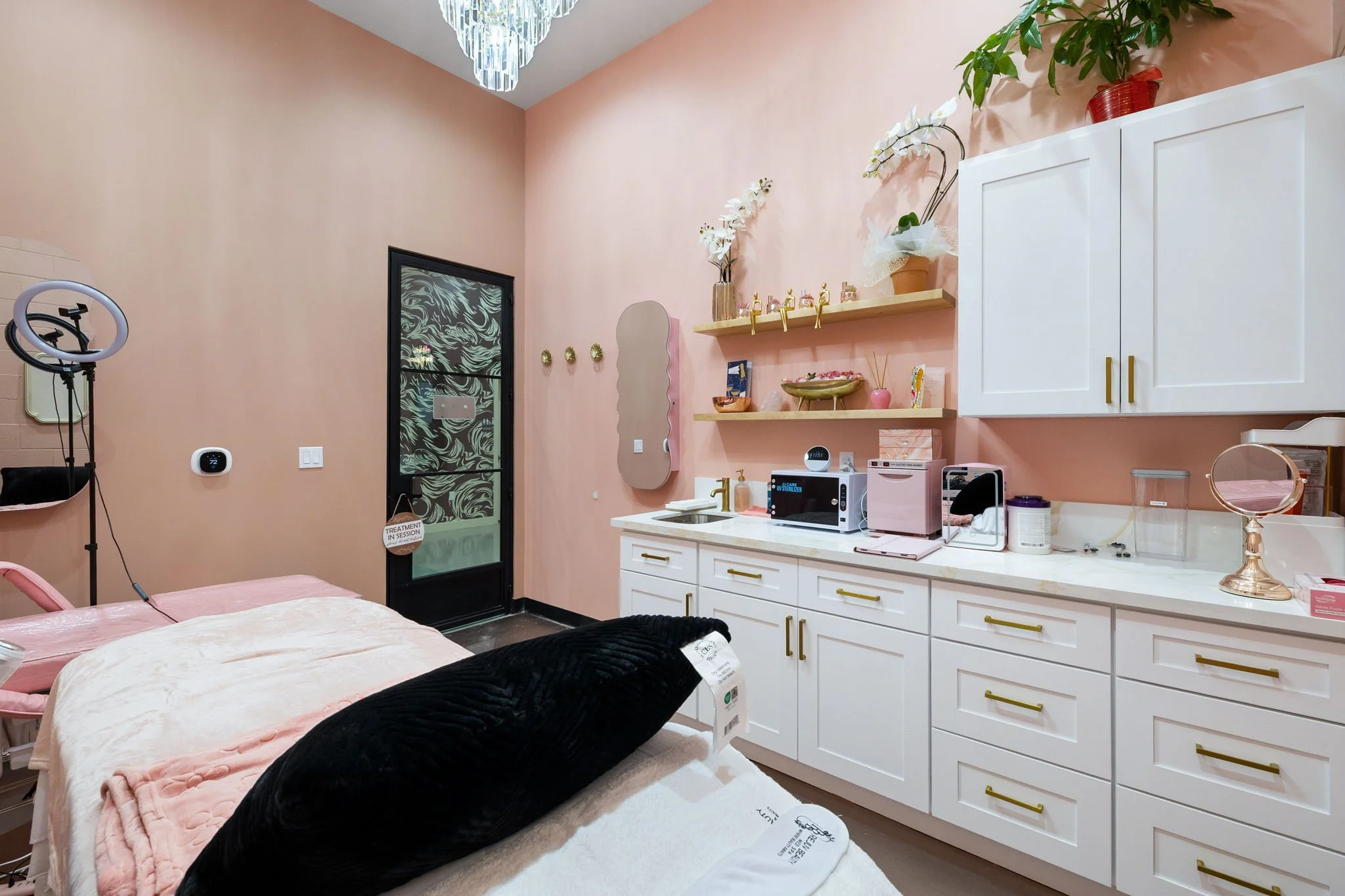 A cozy and aesthetically decorated spa treatment room with soft pink walls, white cabinets with gold handles, and open shelves holding decorative items and plants. There is a treatment bed with a plush black pillow and light pink bedding, a ring ligh