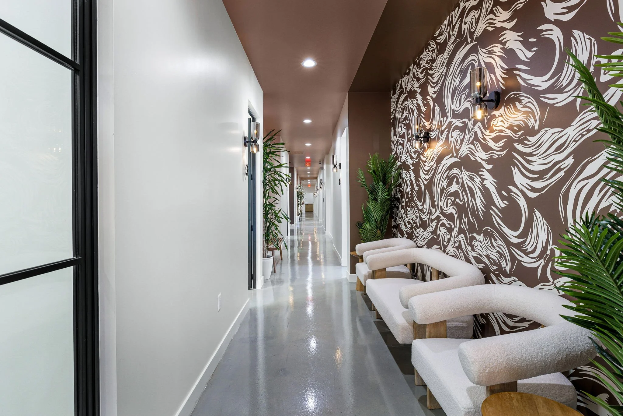 Modern hallway with white walls, decorative brown wall with abstract white swirl pattern, beige curved armchairs, potted plants, wall sconces, and polished gray floor tiles.