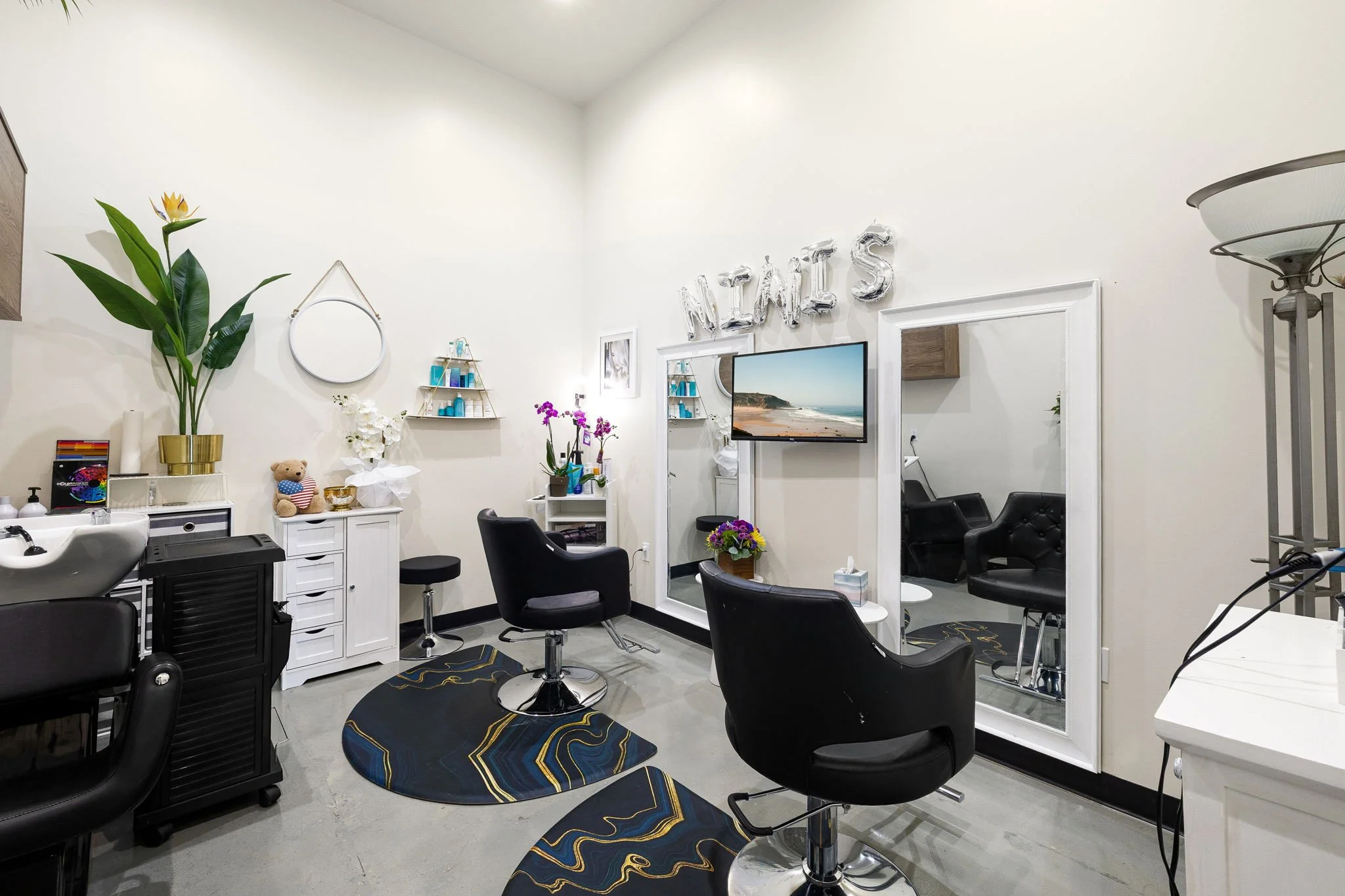 Interior of a hair salon featuring black salon chairs, a mirror, a TV, plants, and decorative balloons spelling 'NAILS' on the wall.