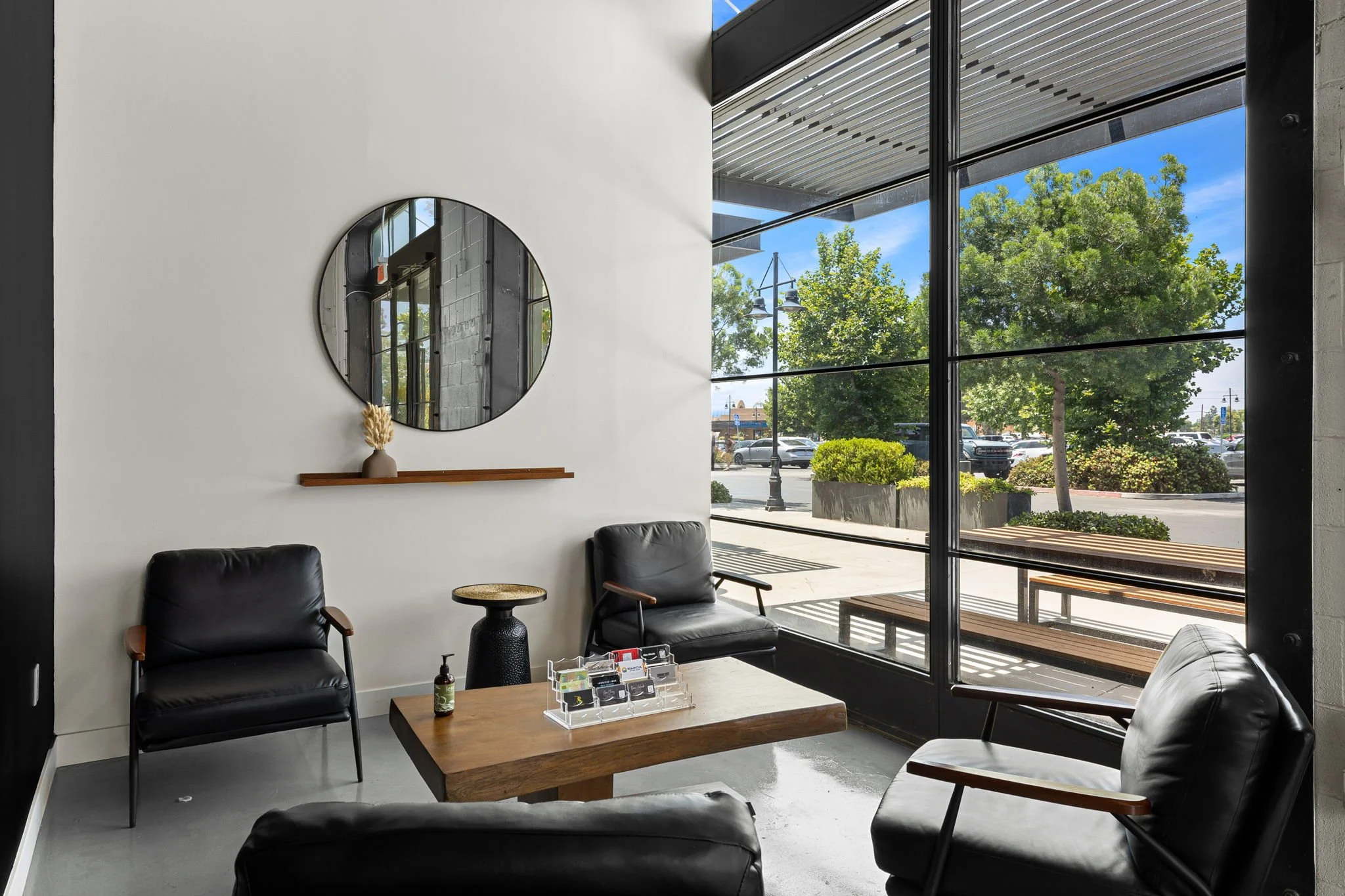 Interior of a modern waiting area with black leather chairs, a wooden coffee table, and a window overlooking a parking lot with trees and benches.