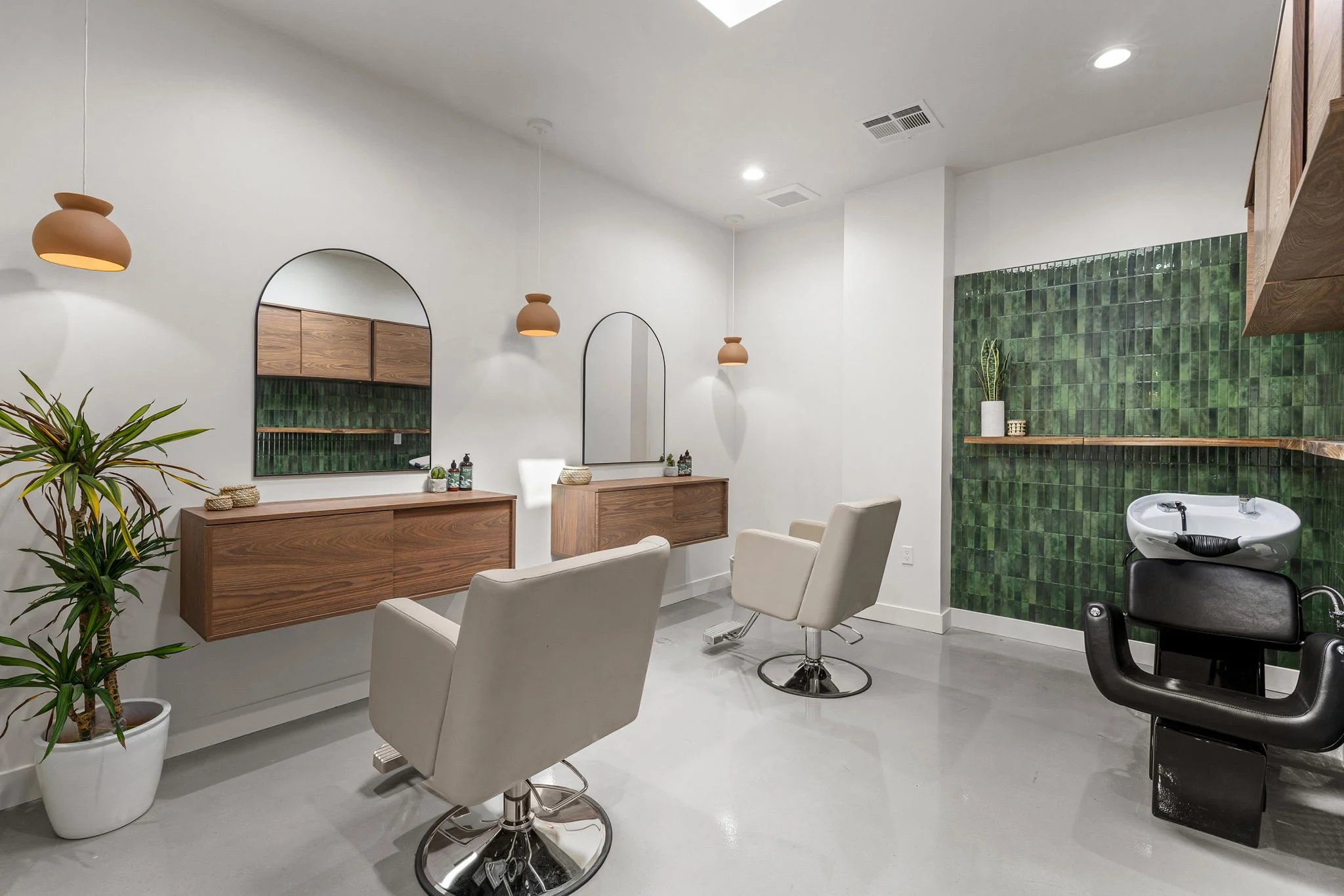 Modern salon with two styling chairs, wall mirrors, wooden shelves, green tiled accent wall, hair washing sink, potted plant, and minimalist decor.