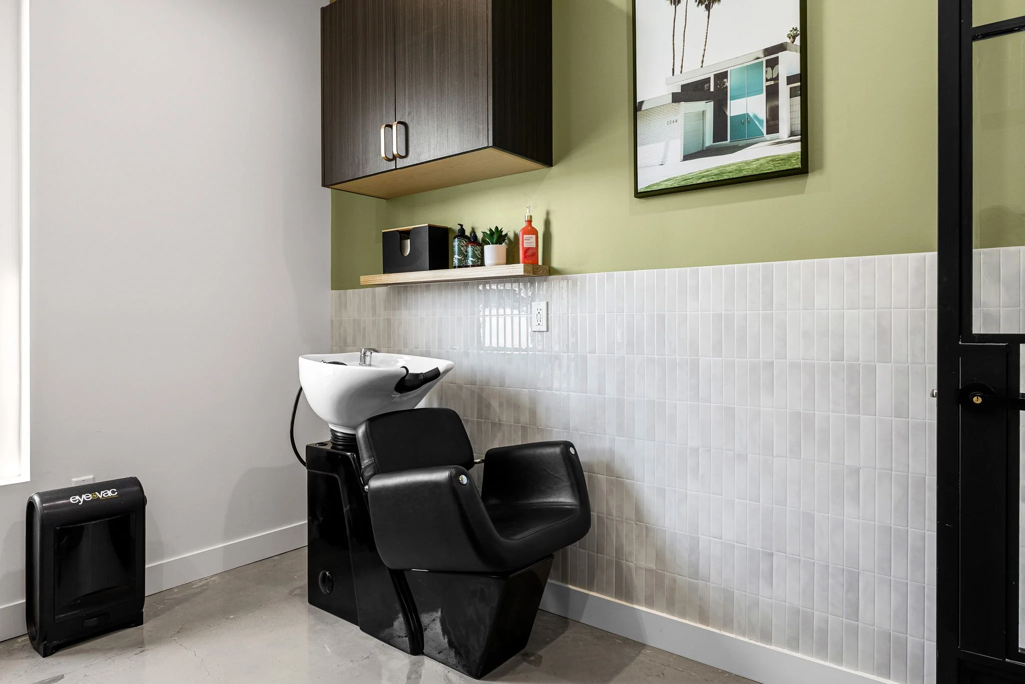 A salon hair washing station with a black chair, white sink and wall-mounted cabinet in a well-lit room.