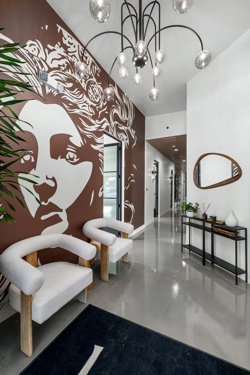 Modern interior hallway with a large wall mural of a woman’s face, white chairs, a black console table with decorative items, a mirror, and a stylish chandelier.