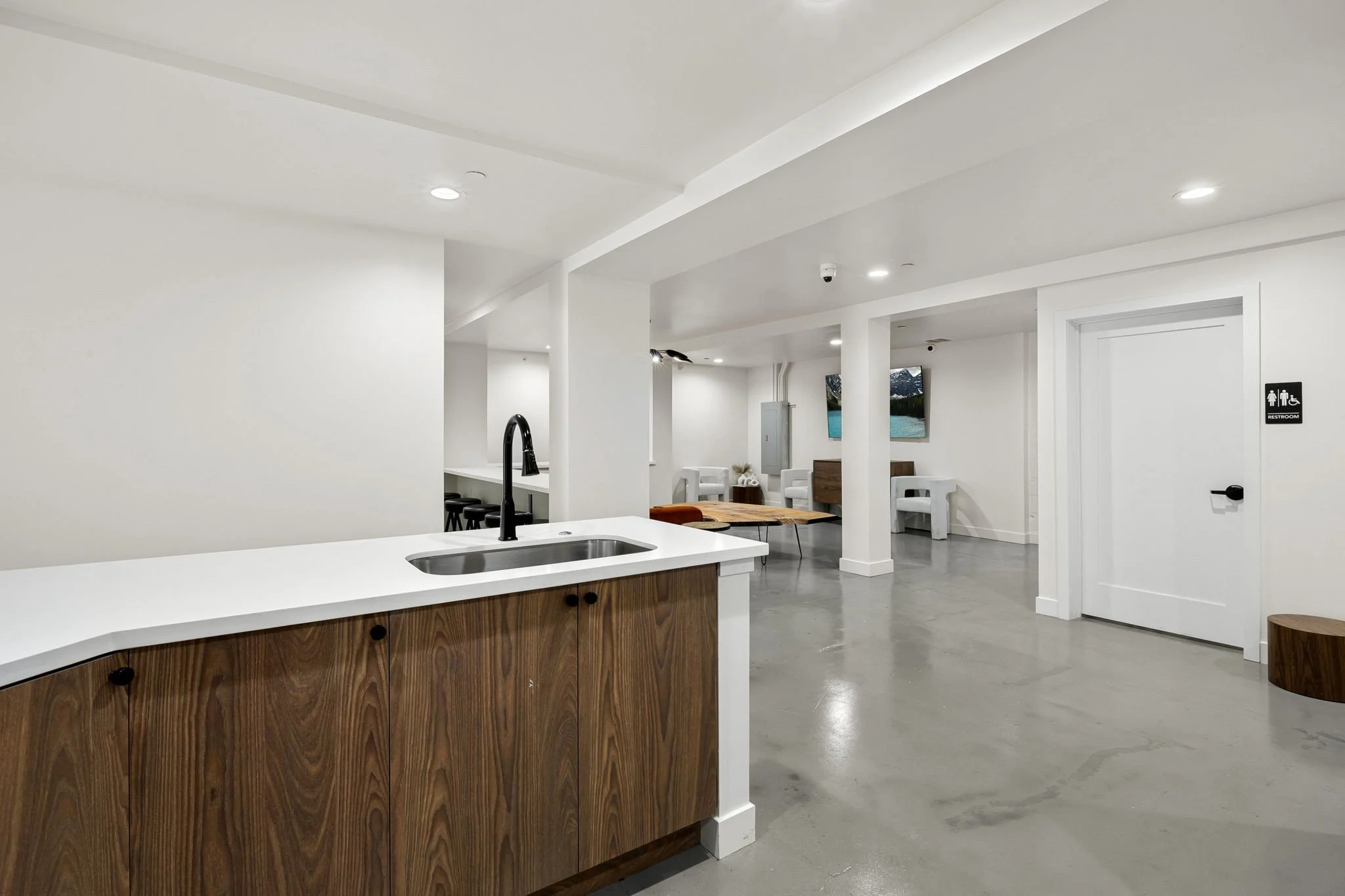 Modern, minimalist interior with a white kitchen counter, stainless steel sink, and black faucet. In the background, there is a lounge area with white chairs, wall-mounted TV screens, and a doorway marked as restroom.