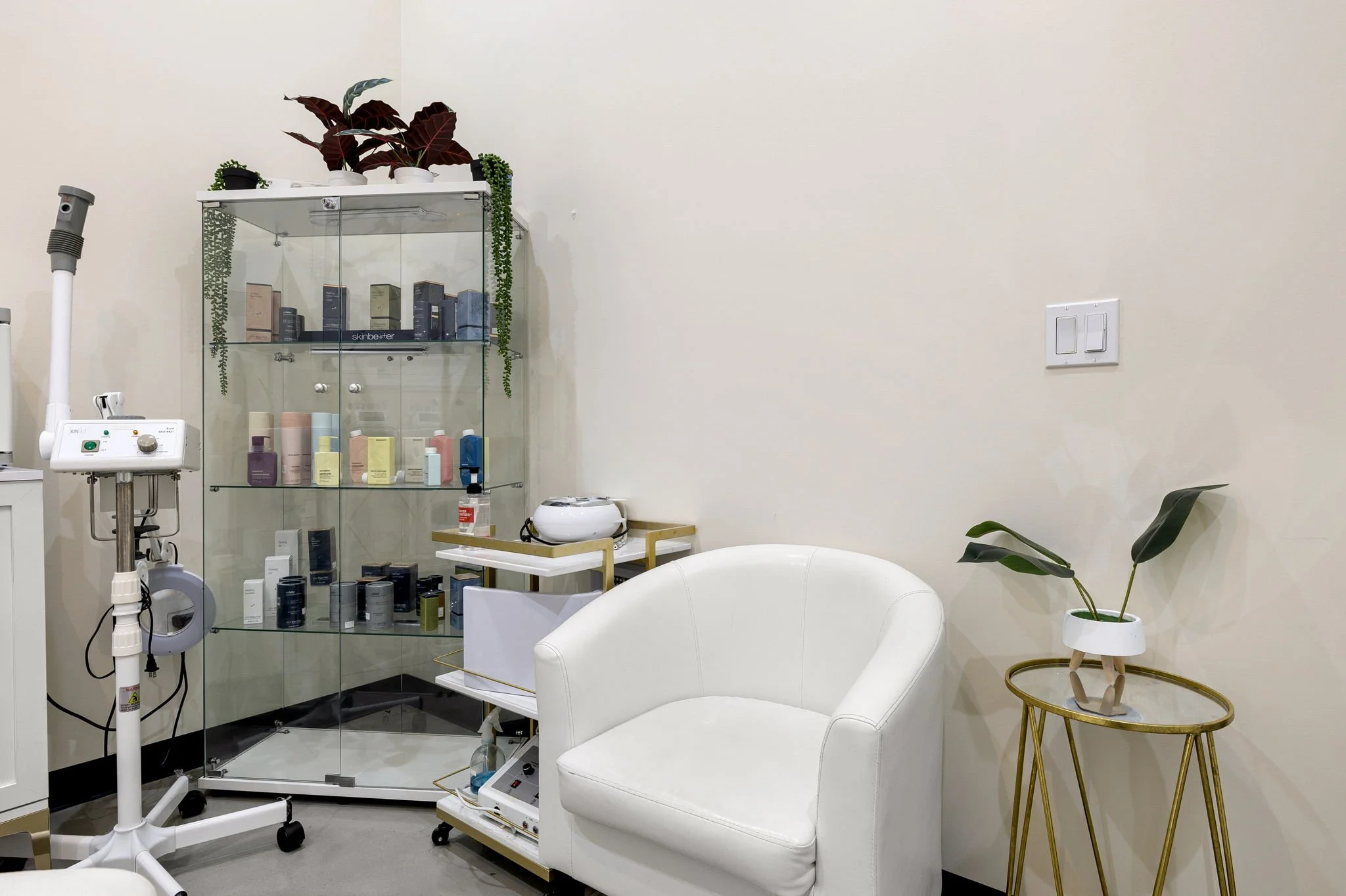 A clean and minimalistic room with a white armchair, a small gold side table with a potted plant, a display cabinet with skincare products, and medical equipment including a lamp and a machine, in a neutral-colored space.