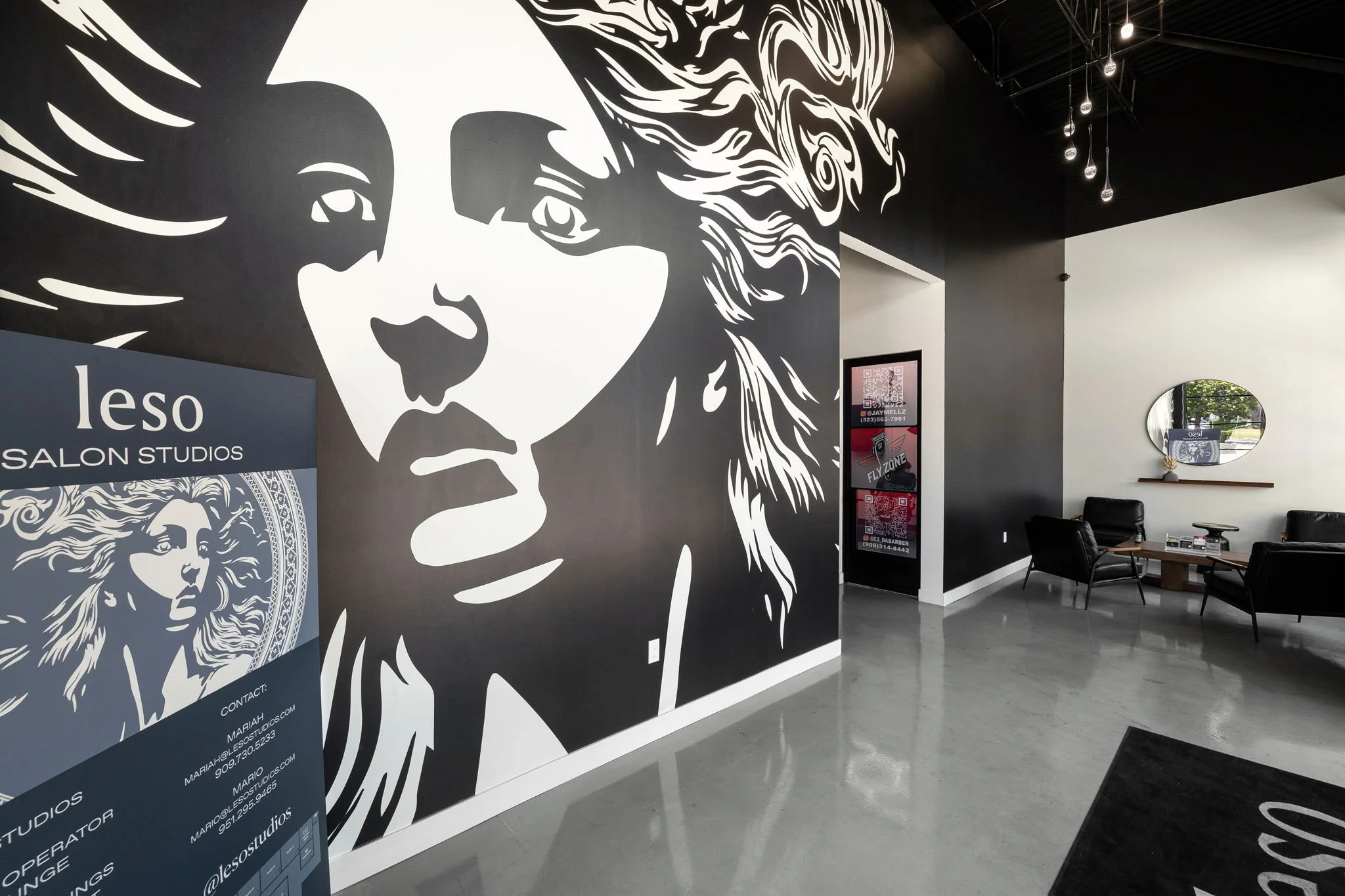 Interior of a salon or studio with a large black and white mural of a woman's face on the wall, black chairs, a mirror on the white wall, and informational panels near the entrance.