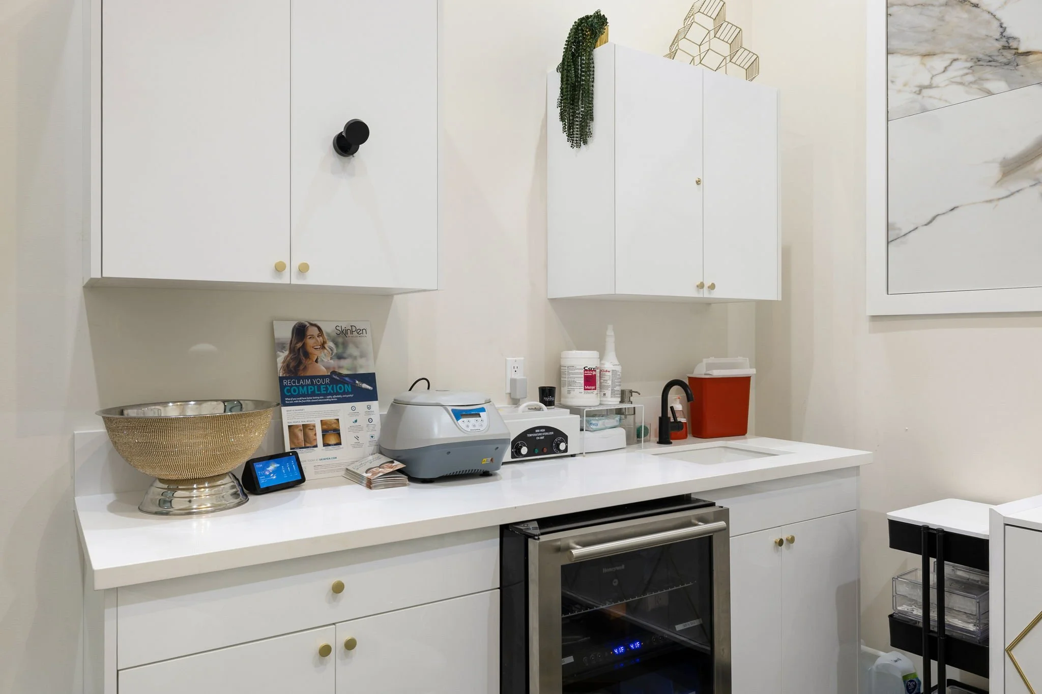 A medical or laboratory room with white cabinets, a countertop with various equipment, a small sink, and a wine cooler, with medical supplies and informational brochures on the counter.
