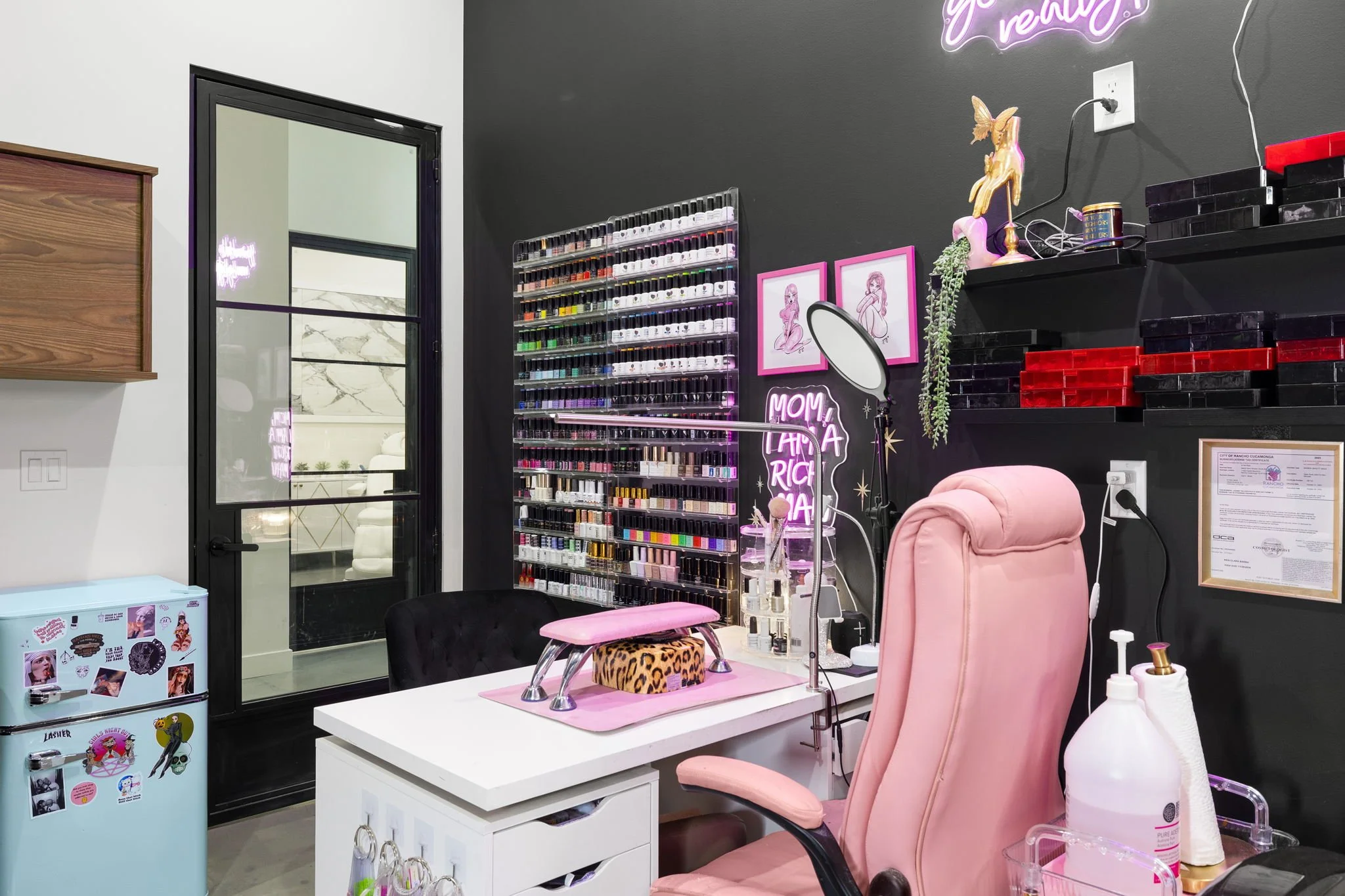 Interior of a nail salon or beauty spa with a pink and black color scheme, featuring nail polish displays, a manicure table, pink chair, decorative wall art, and shelves with organized supplies.