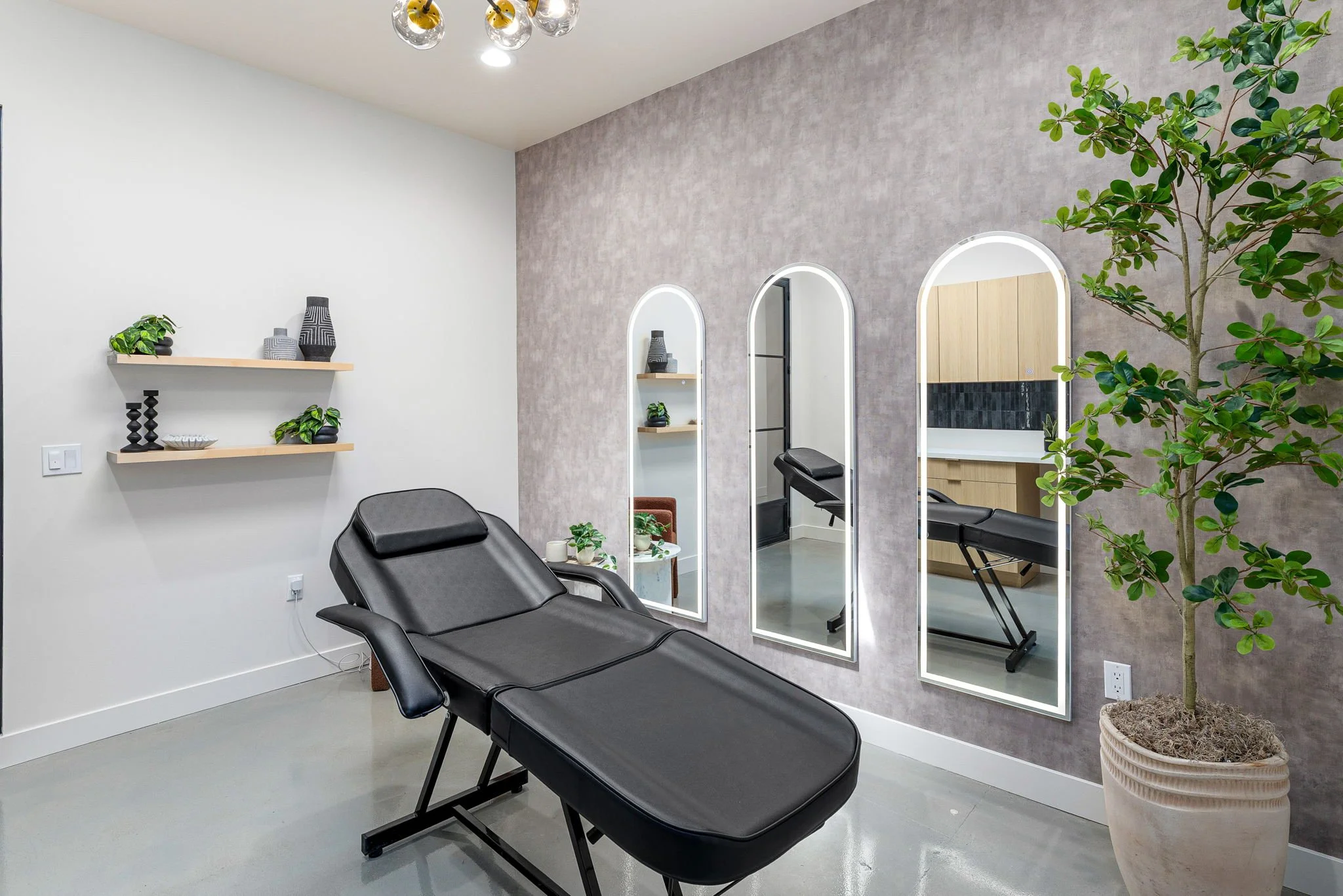 Interior of a spa or massage therapy room with a black adjustable treatment table, three tall white-framed mirrors on a textured gray wall, a potted plant, and minimal decorative shelves.