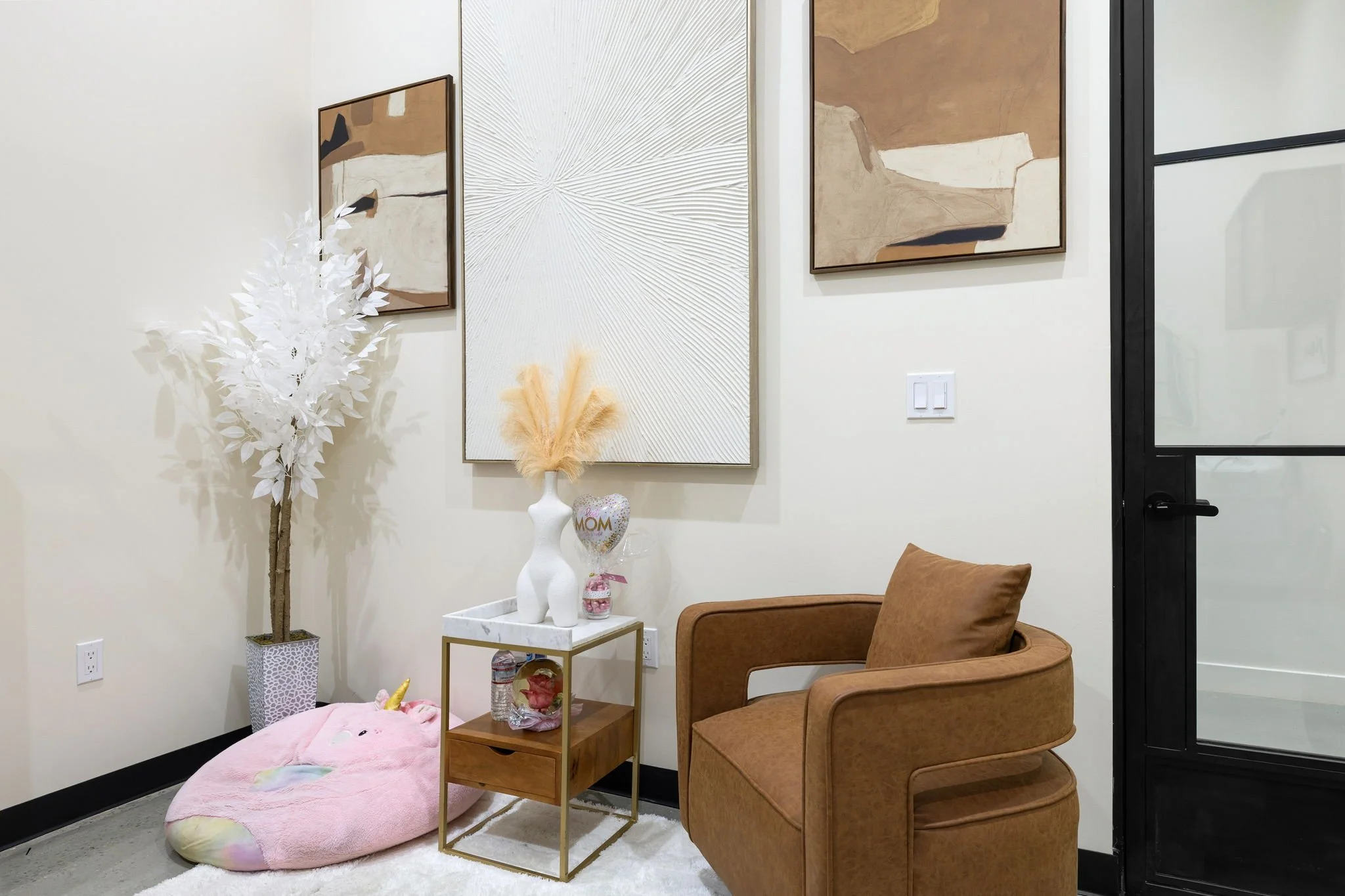 Interior of a modern waiting area with a tan armchair, side table, decorative art on the walls, a white artificial tree, and a pink plush unicorn rug.
