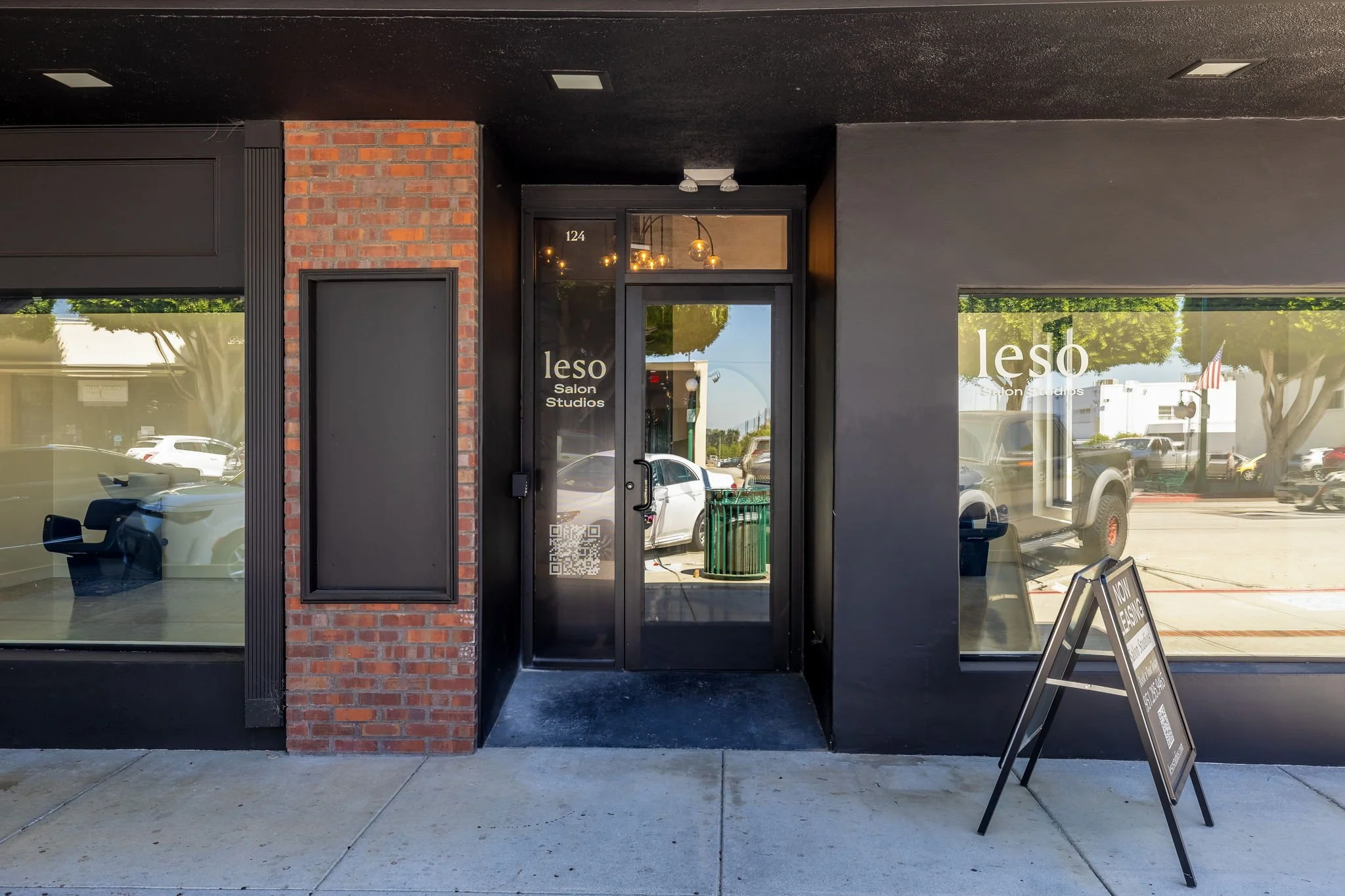 Storefront entrance with black walls and windows, a red brick pillar inside the sidewalk, and signage displaying 'leso Salon Studios'