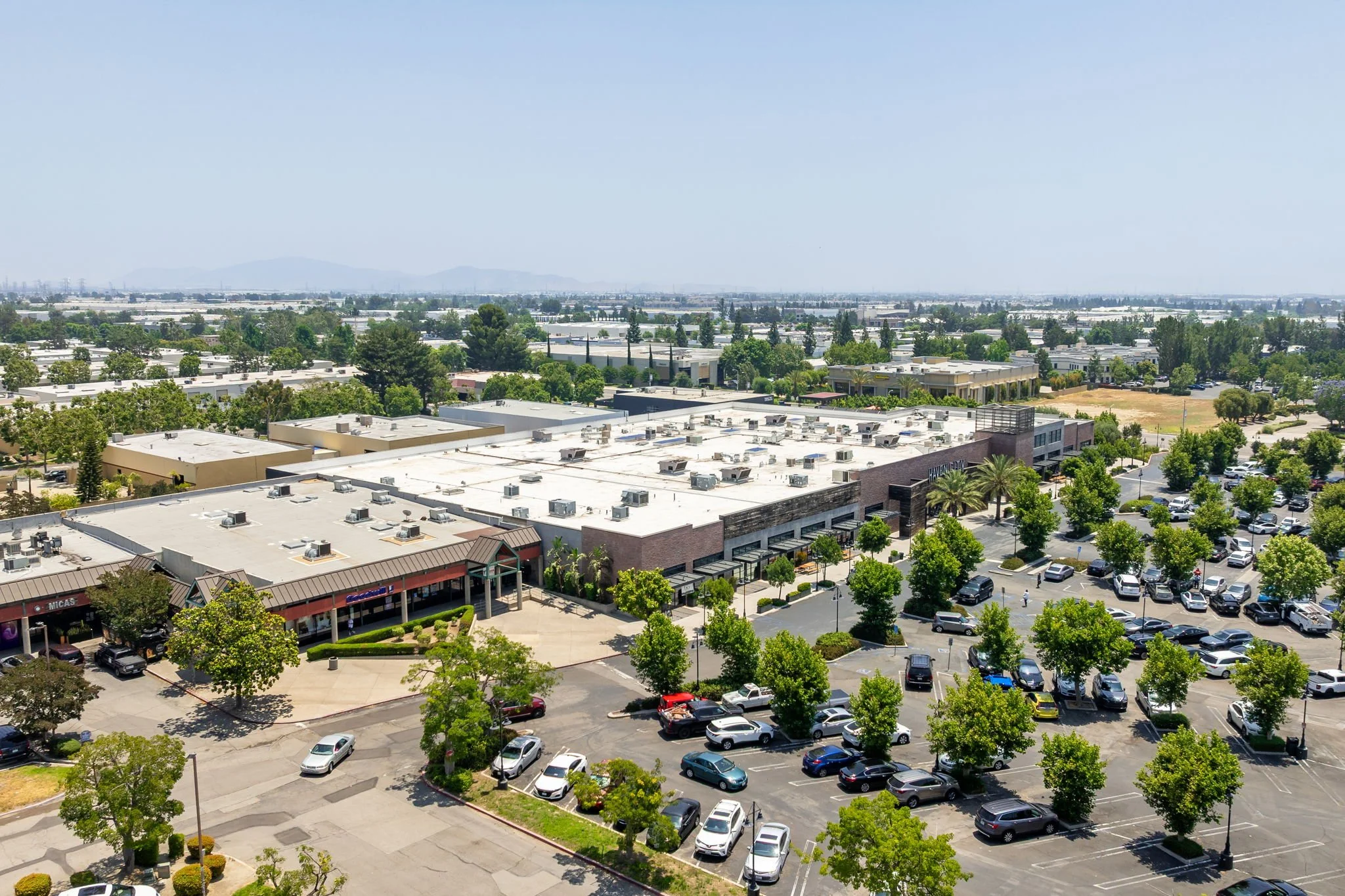 Airy view of a shopping plaza and adjacent parking lot filled with cars and surrounded by green trees, with a large commercial building and distant landscape in the background.