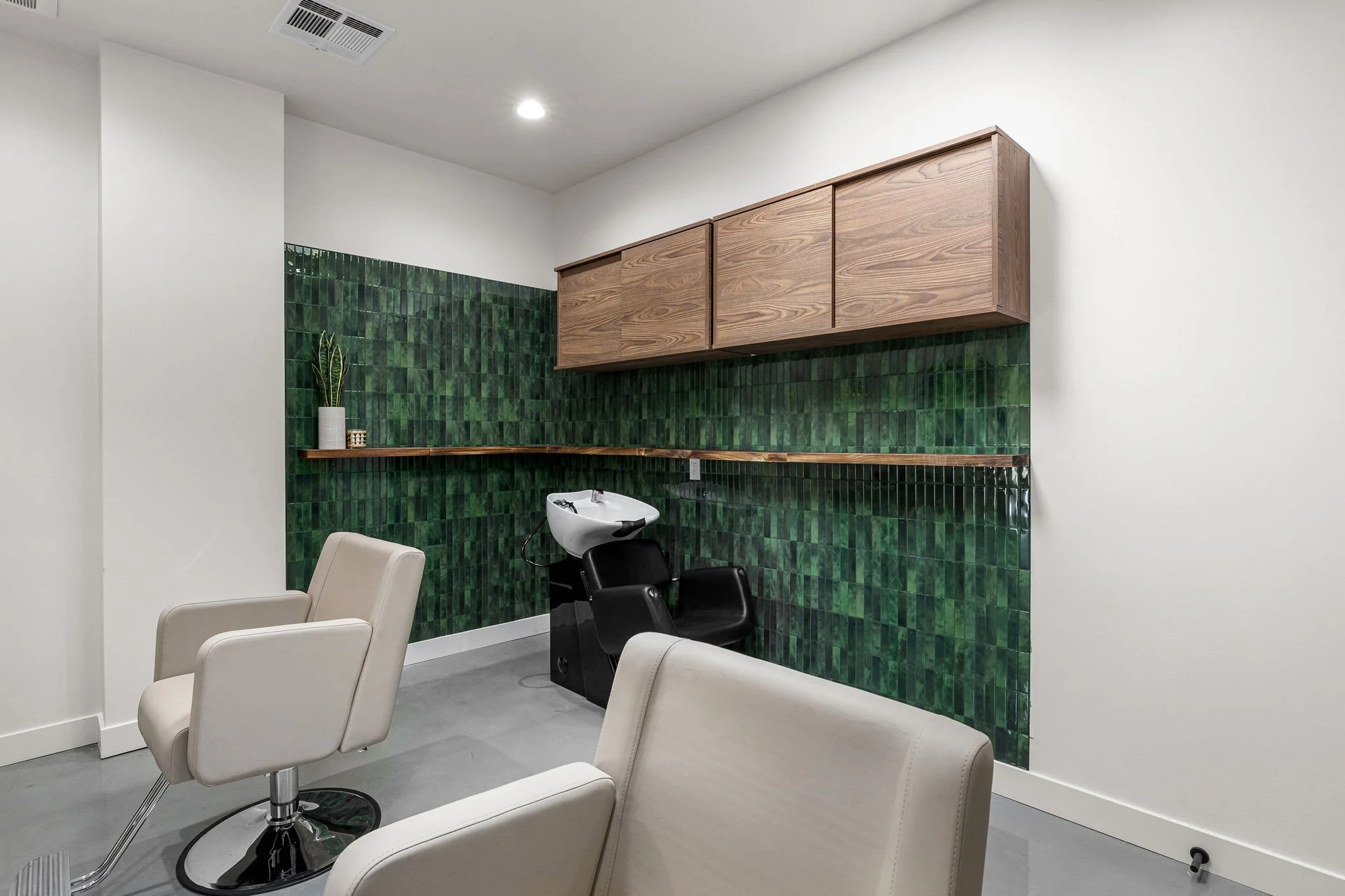 Interior of a modern hair salon or beauty salon showcasing a white styling chair, a hair washing basin, and green tiled wall with wooden cabinets and a shelf with a plant on it.