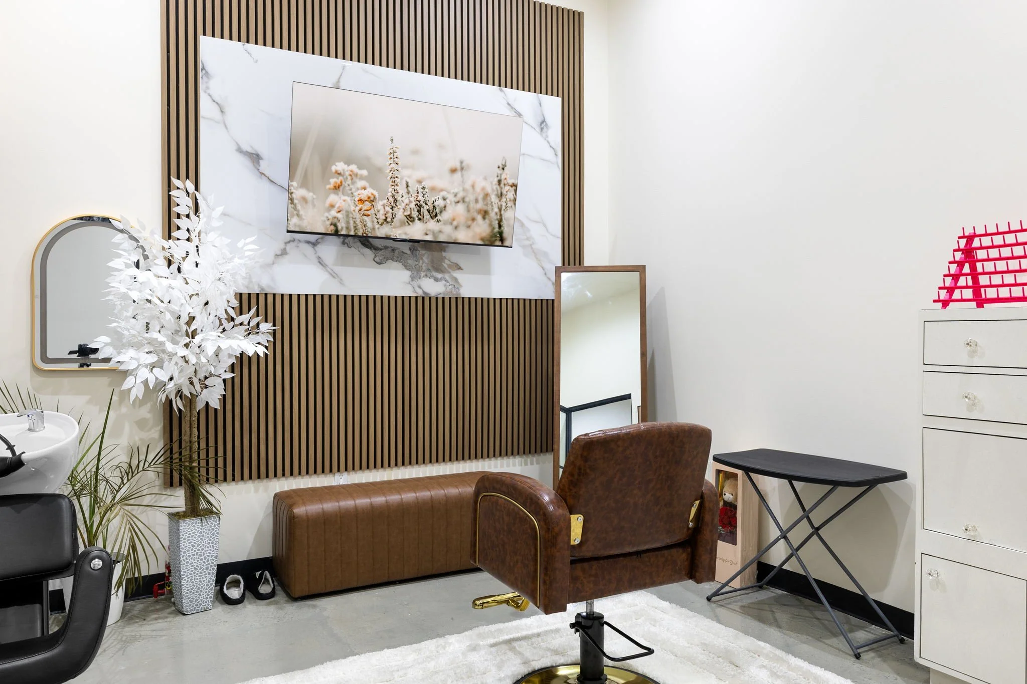 Stylish salon interior with a leather barber chair, a mirror, a TV mounted on a marble-patterned wall, a white decorative tree, a cushioned bench, and a dresser with a pink tray on top.