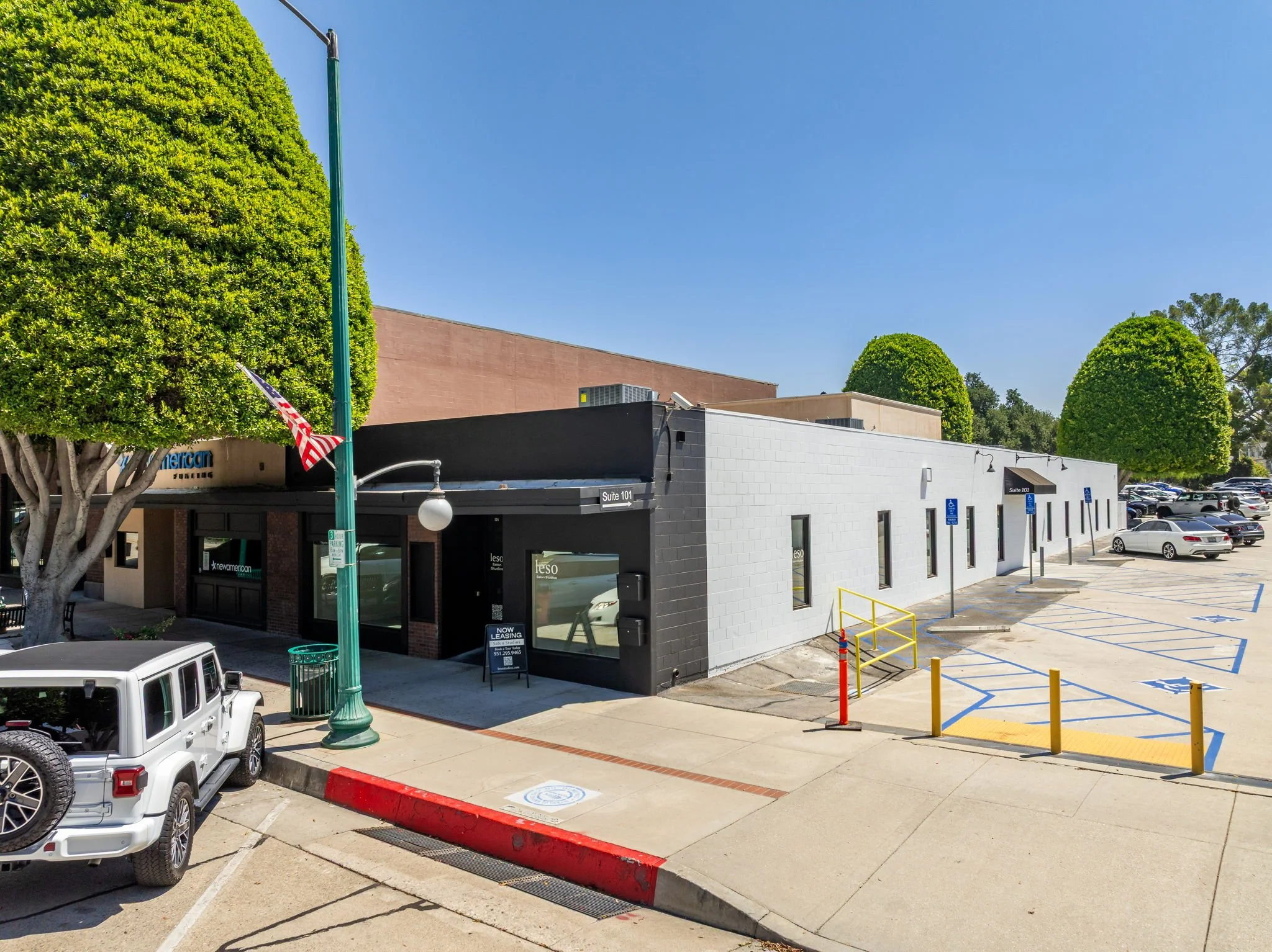 A parking lot in front of a commercial building with trees, parked cars, and corner ramps for disabled access.