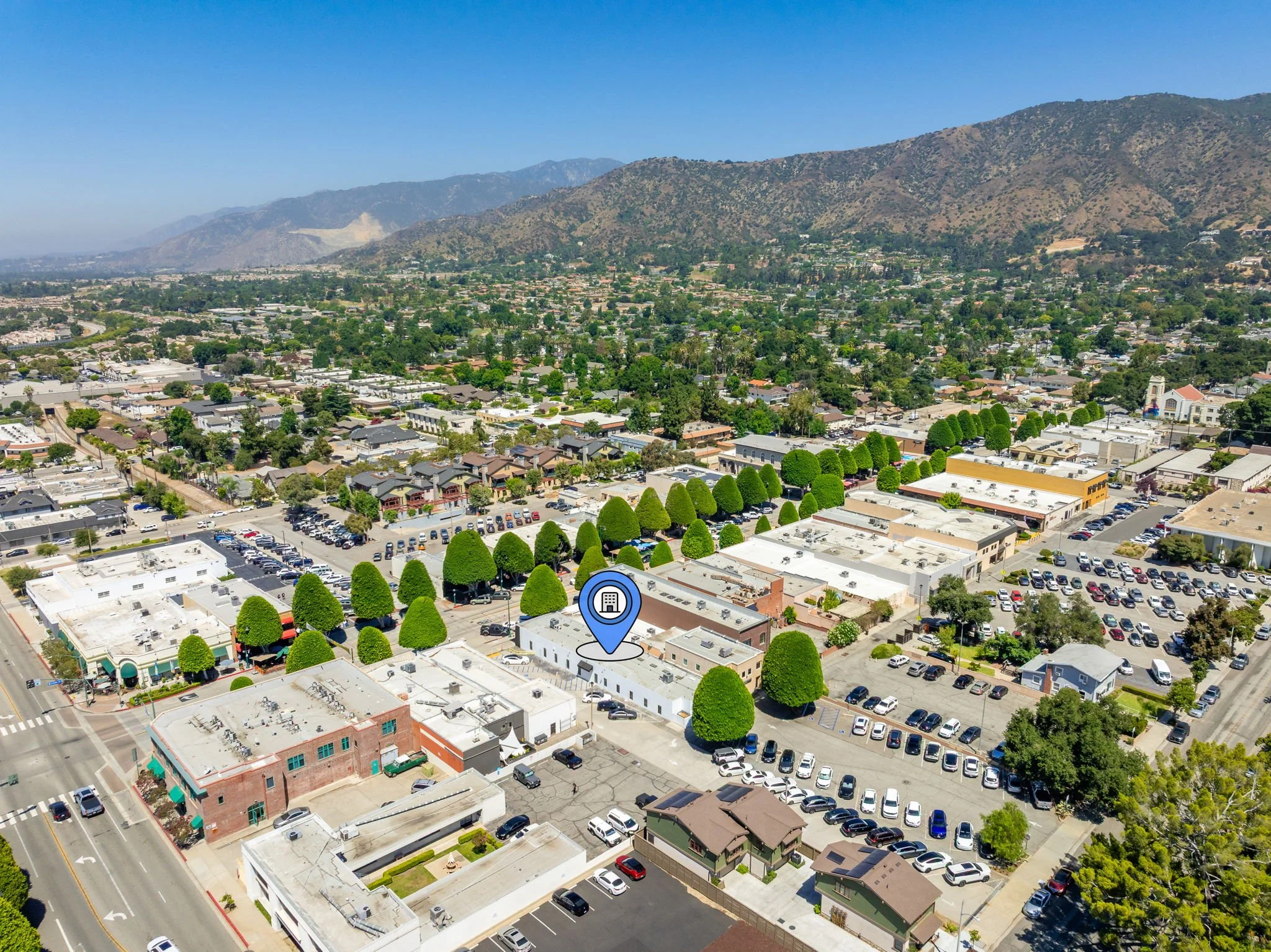 Aerial view of a commercial area with parking lots, buildings, and trees, with a mountain range in the background.
