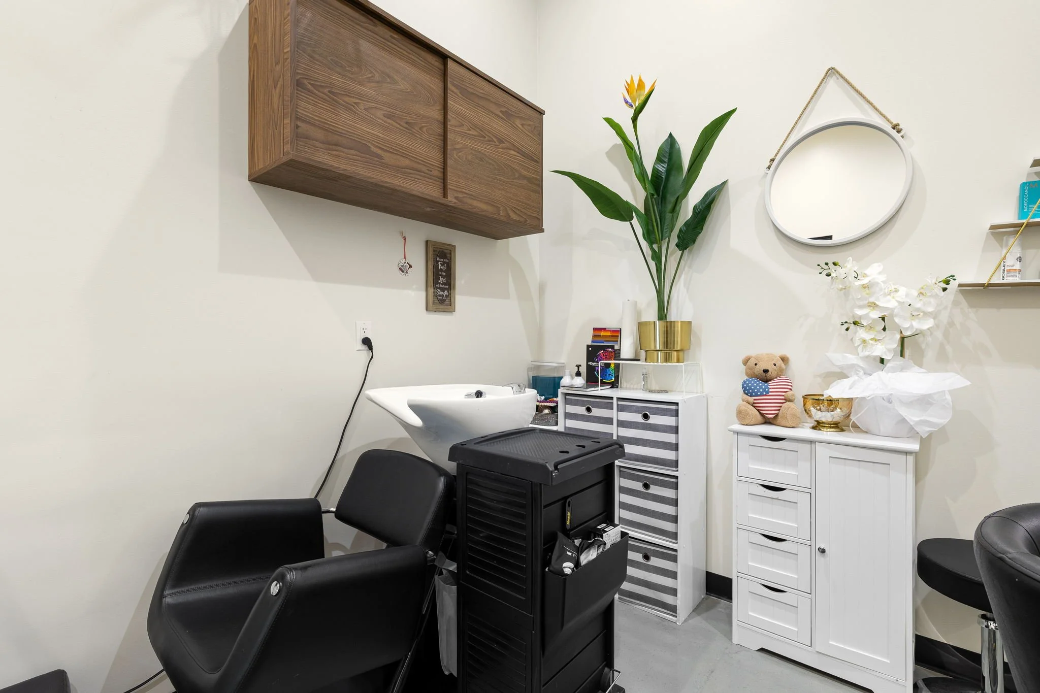 Hair salon with a black styling chair, a white sink, a striped storage cabinet, decorative plants, a teddy bear, a mirror, and various hair tools.