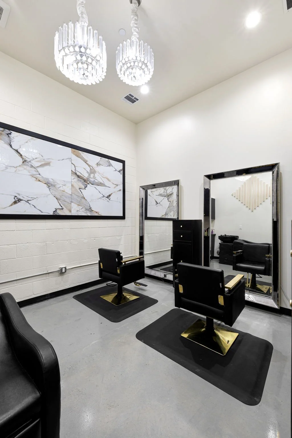 Interior of a modern hair salon with black styling chairs, large mirrors, a marble wall art piece, and a chandelier on a white ceiling.
