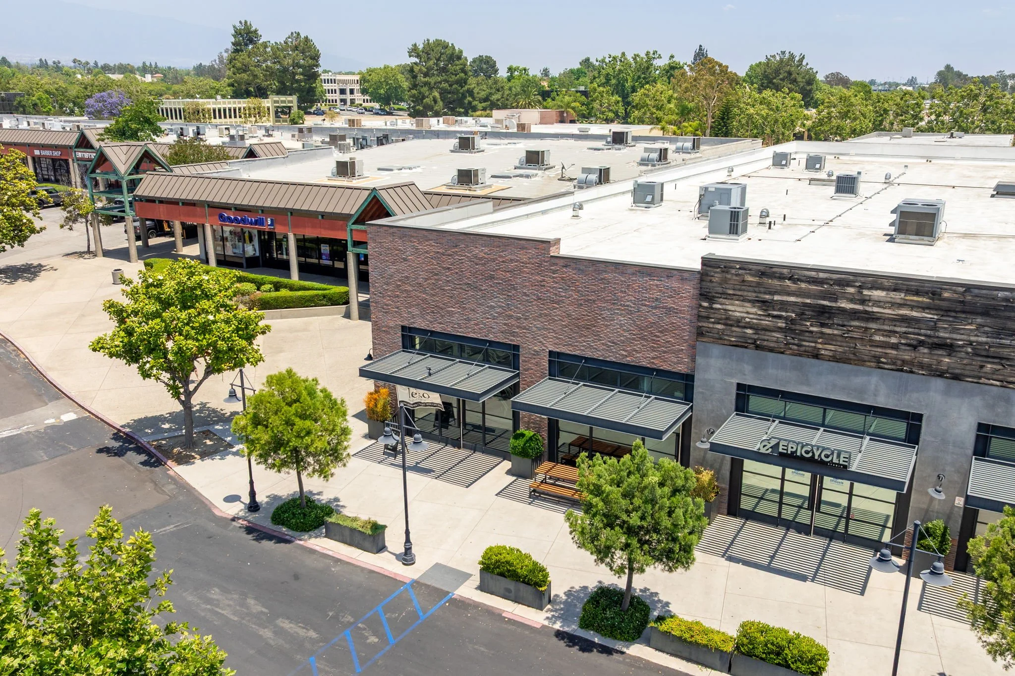An aerial view of a shopping center with retail stores, trees, parking lot, and a large white roof with multiple HVAC units.