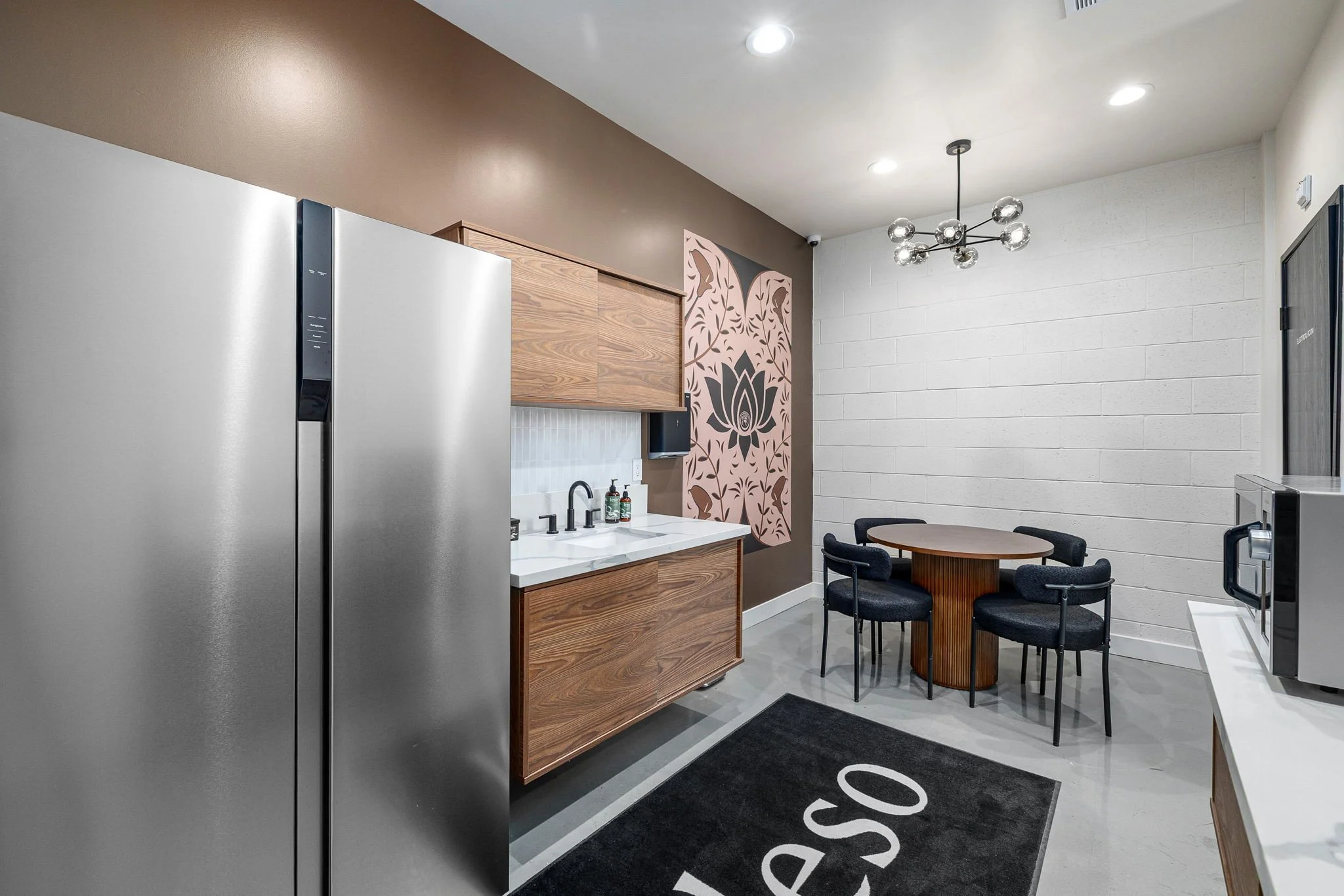 Modern kitchen and dining area with stainless steel refrigerator, wooden cabinets, black sink, small round dining table with four black chairs, decorative wall art, and a black rug with white letters.