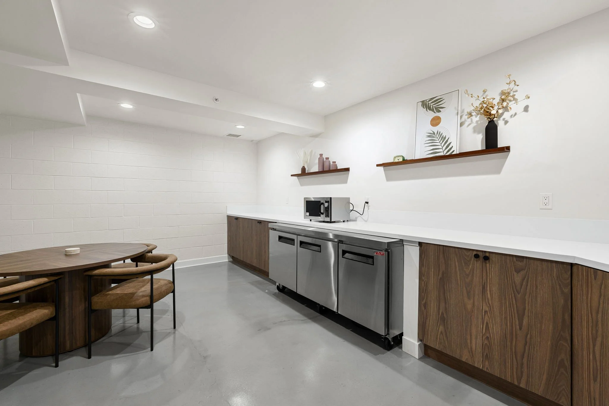 Modern kitchenette with white countertops, wooden cabinets, stainless steel mini fridge, microwave, and decorative shelves with vases and framed art, adjacent to a wooden dining table and chairs.