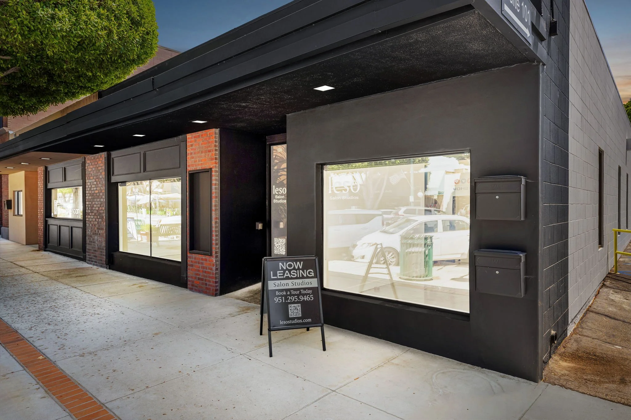 Exterior view of a black building with large glass windows, a sidewalk in front, and a sidewalk sign advertising leasing for salon studios. There are mailboxes on the wall and a parked car visible through the windows.